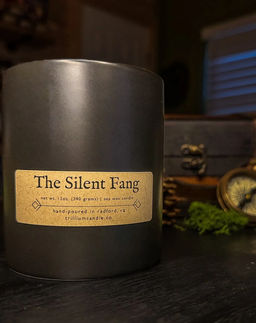 A black soy wax candle with a beige label titled 'The Silent Fang.' The label states it is 12 oz, hand-poured in Radford, VA, and from trilliumcandle.co.