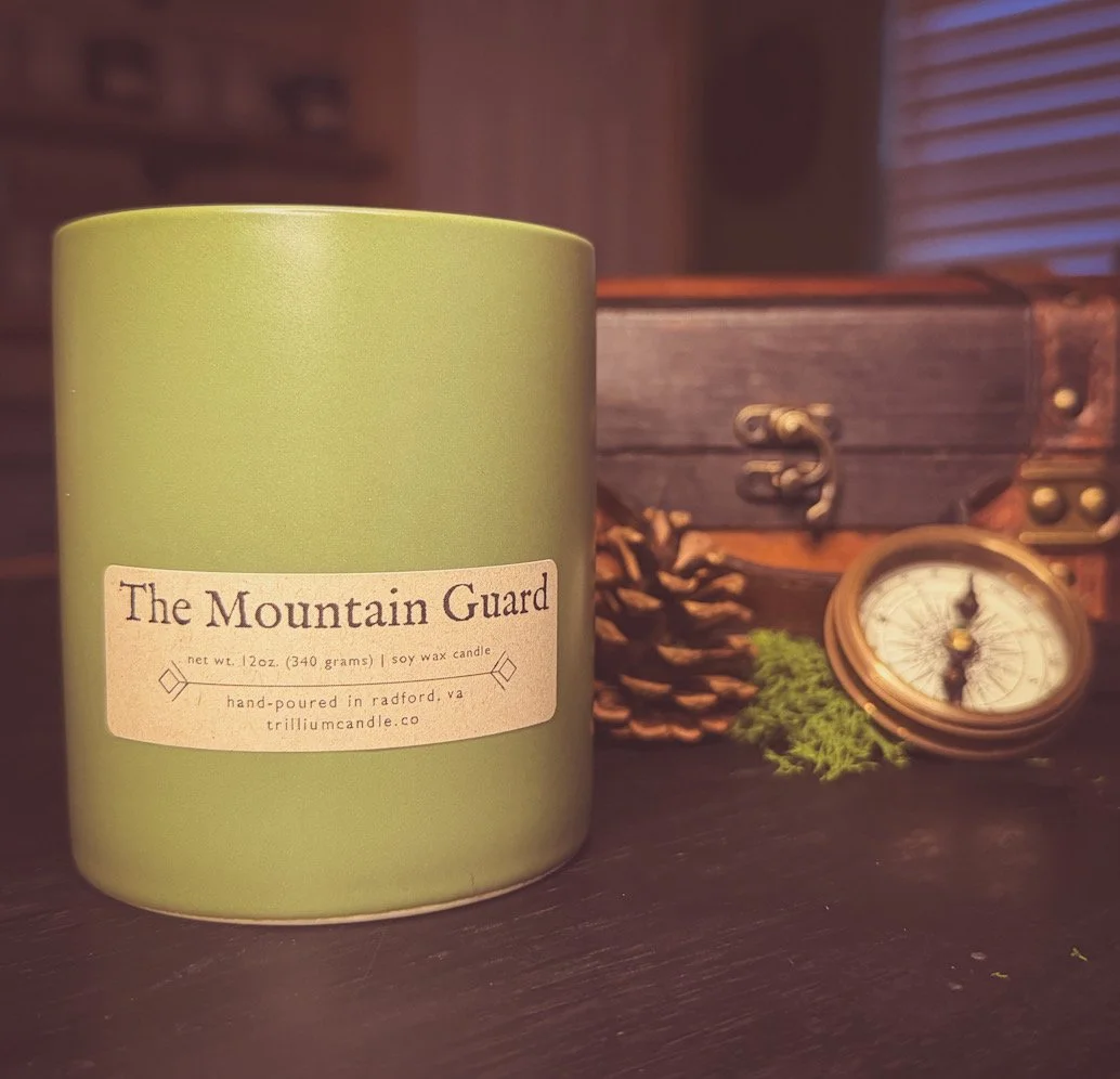 A green candle labeled 'The Mountain Guard' placed on a dark surface, with a pine cone, green moss, a small round compass, and a wooden chest in the background.