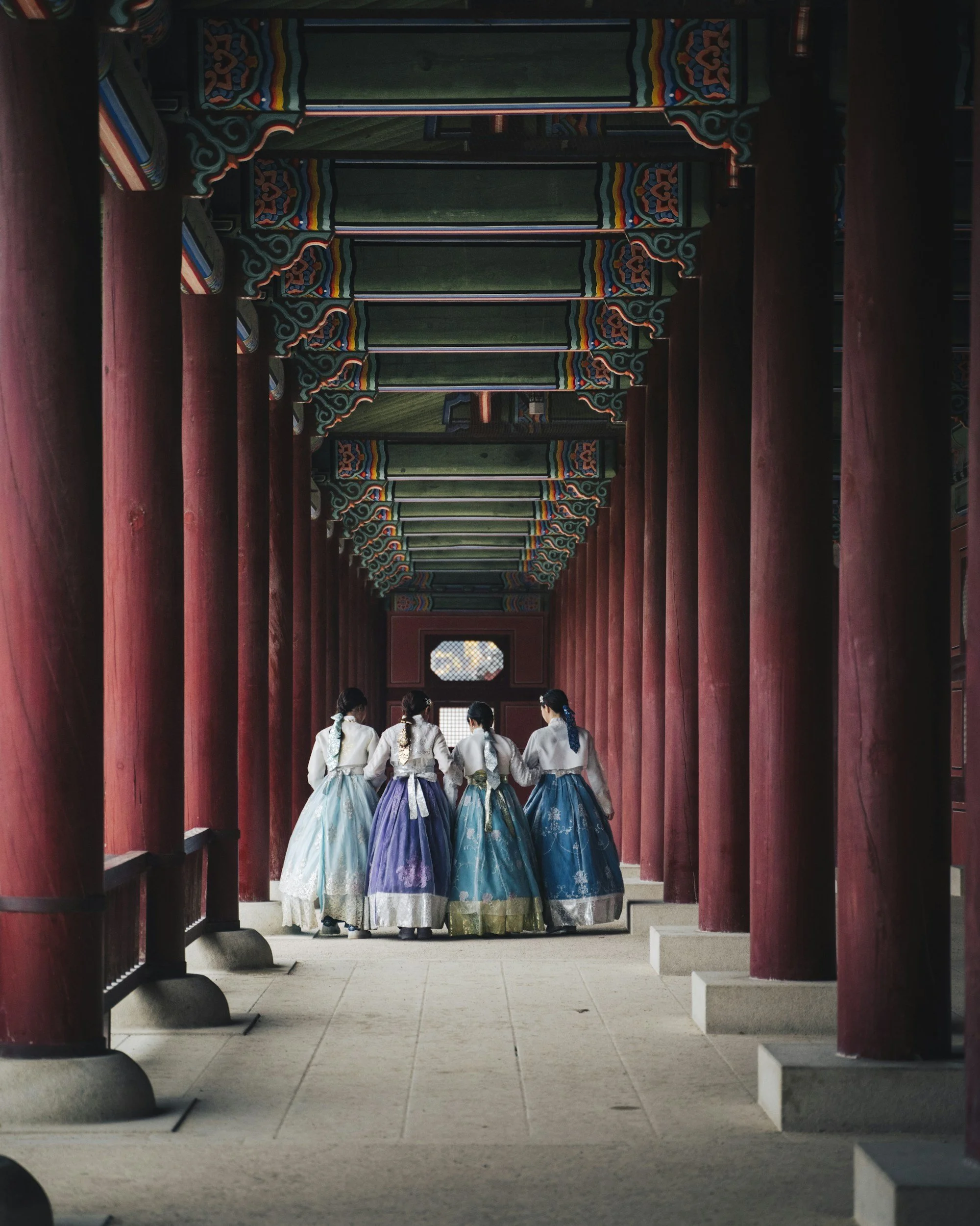 Five women dressed in traditional Korean hanbok walking through a corridor of a historic building with wooden pillars and ornate painted ceiling.