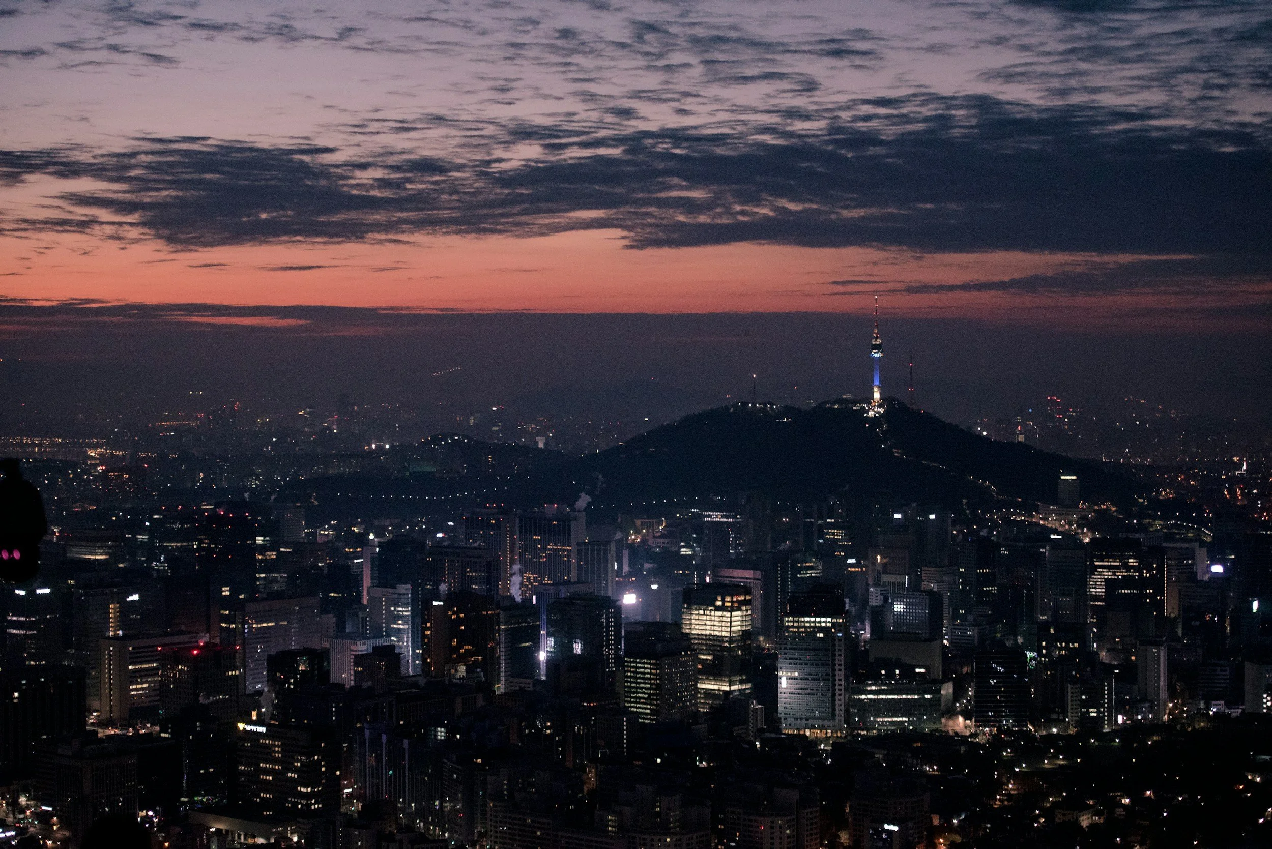 Nighttime cityscape view of Seoul, South Korea, with Namsan Tower illuminated on a hill and a sunset sky in the background.