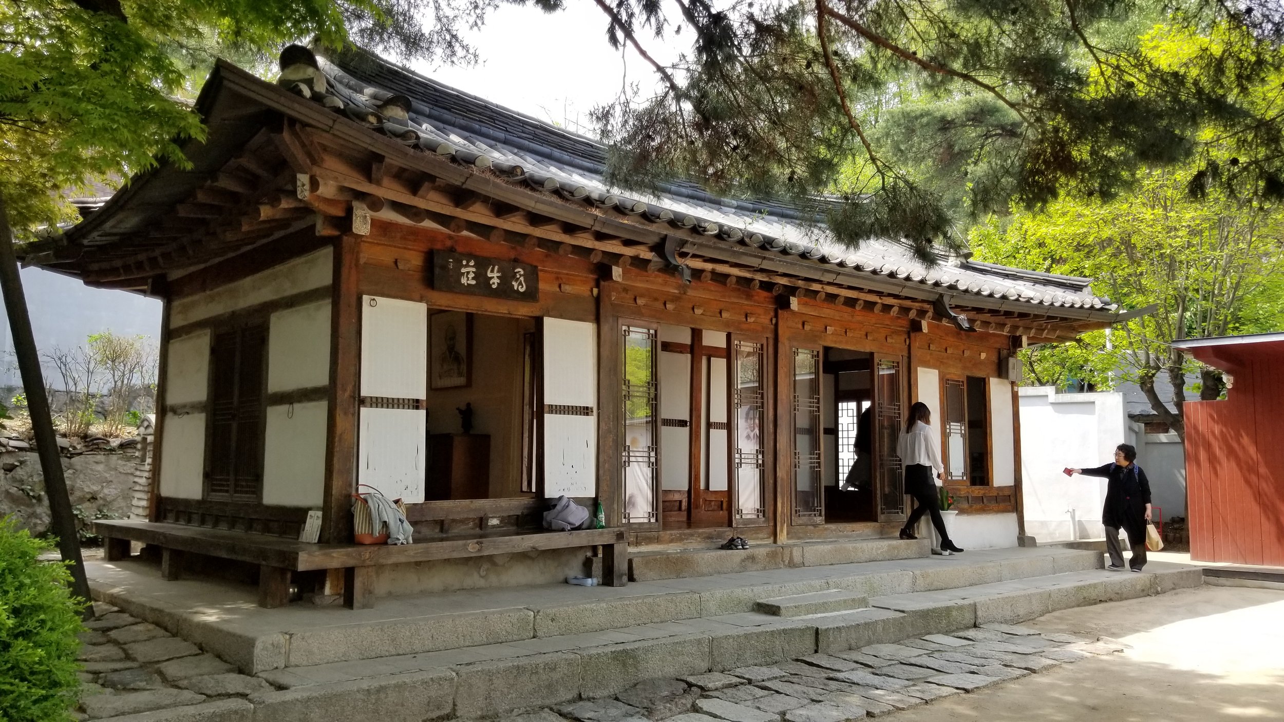 A historical Korean building with a Korea woman pointing at something interesting, there are beautiful trees and plants around the building