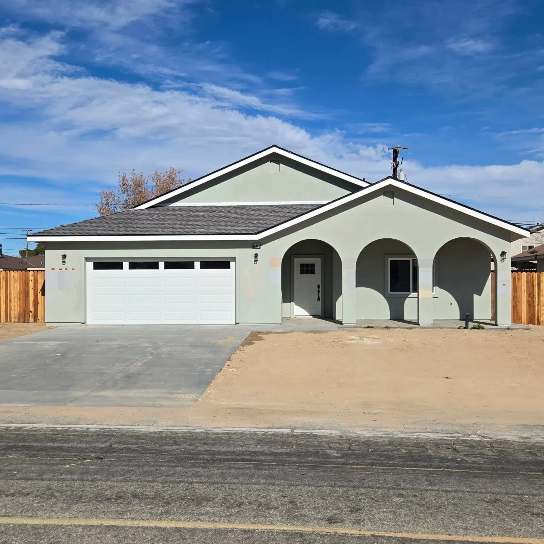 Newly built light green house with a two-car garage, front entrance with three arches, and a concrete driveway, fenced yard, under a partially cloudy blue sky.