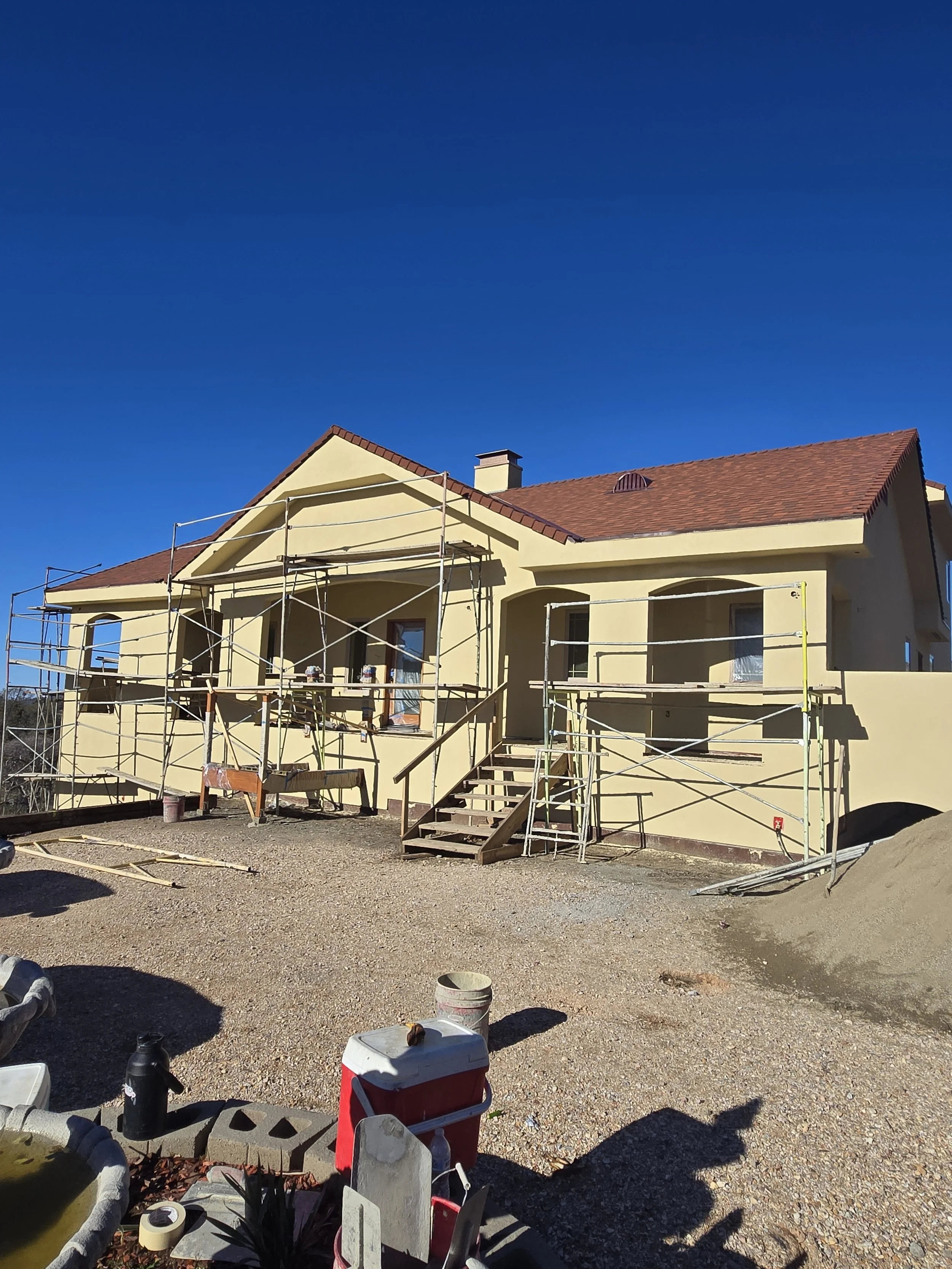 A house under construction with scaffolding around it, a set of stairs leading to the front door, and construction materials in the yard on a clear, sunny day.
