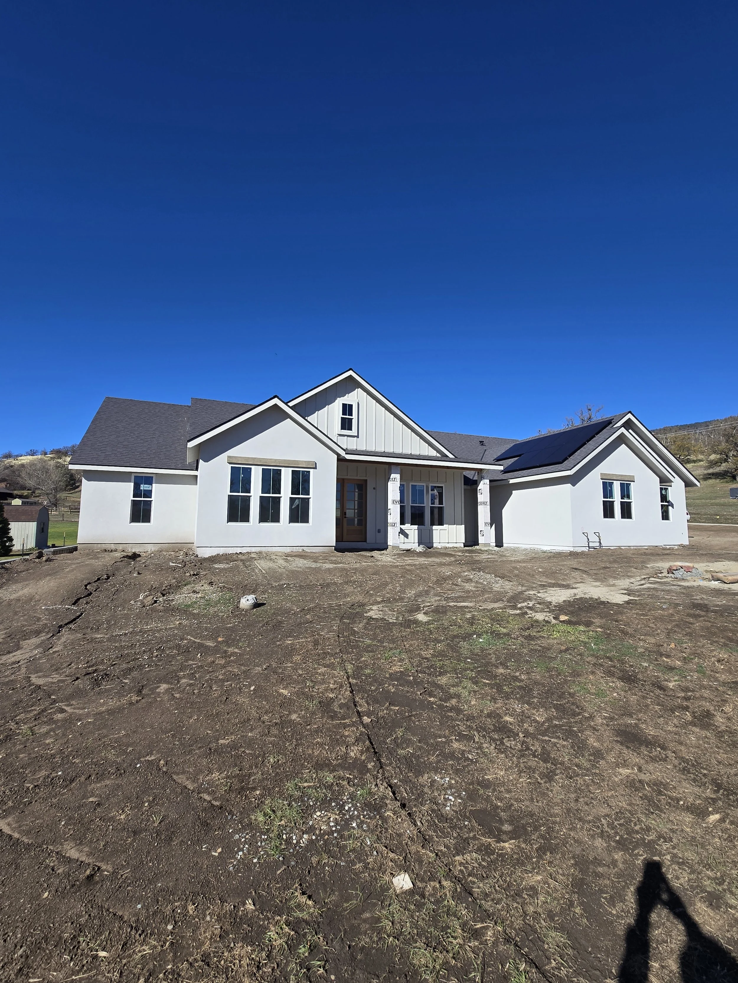 Newly constructed white house with multiple gables, large front windows, and some solar panels on the roof, set against a clear blue sky, with a dirt yard in the foreground.
