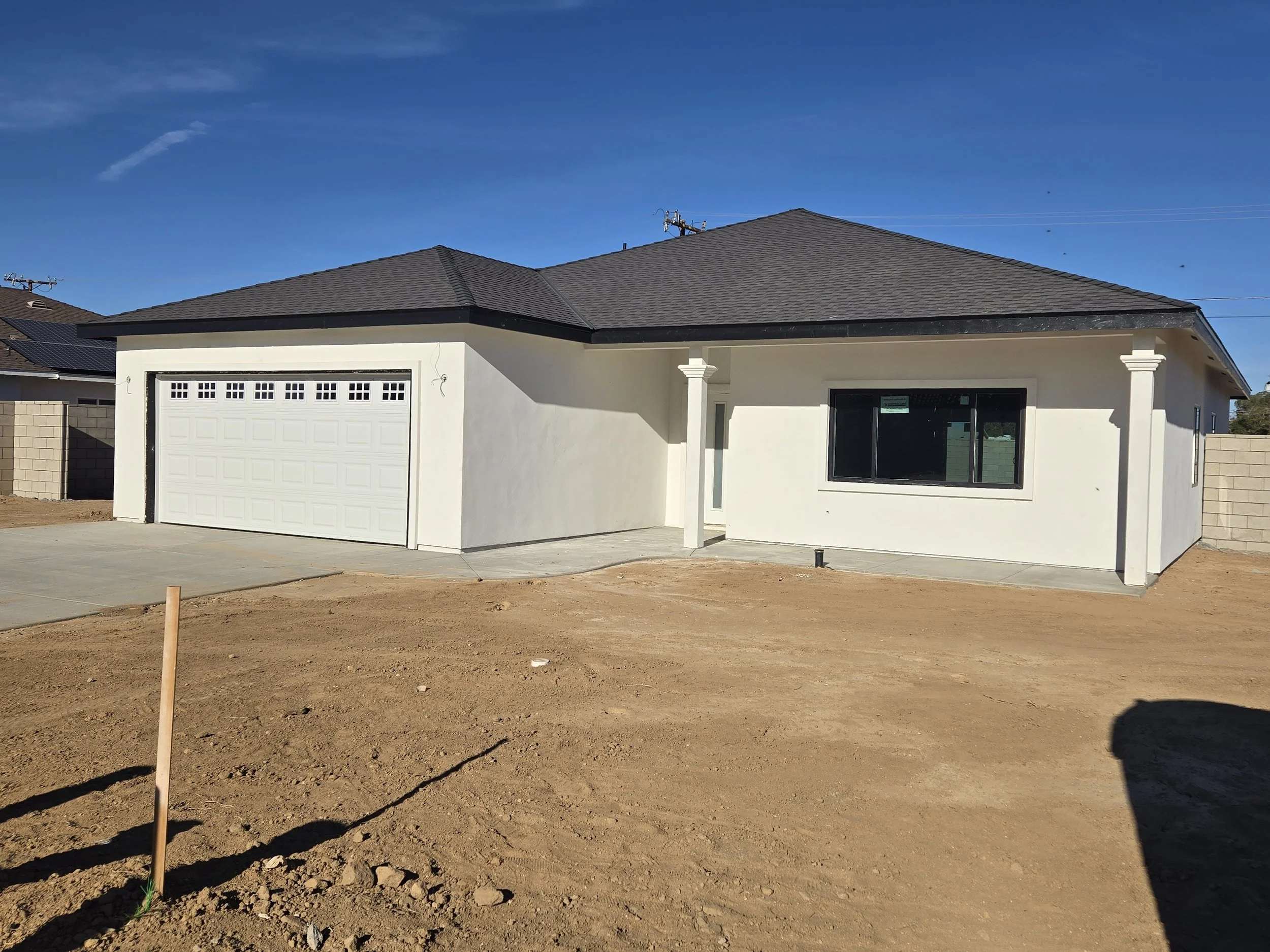 Newly built single-story house with white exterior and black roof, featuring a single-car garage and a large window in a sunny environment, with unpaved yard in front.