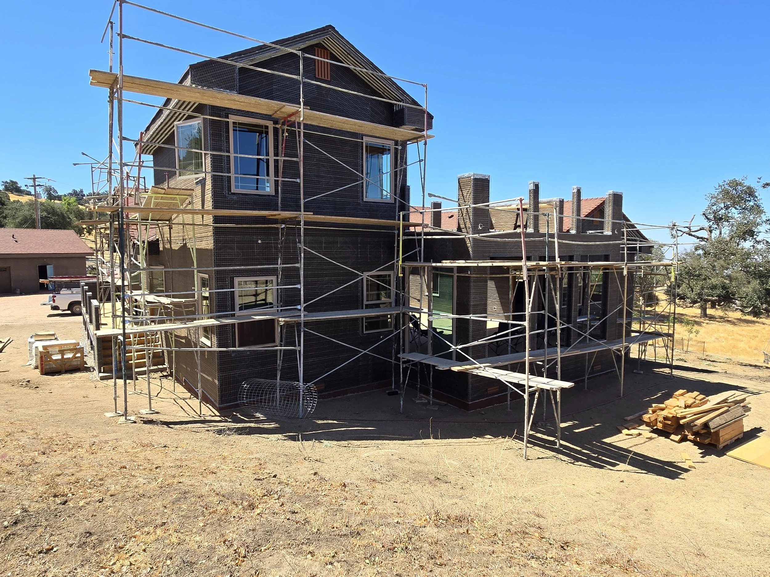 Under construction house with scaffolding on a sunny day, surrounded by dry land and construction materials.