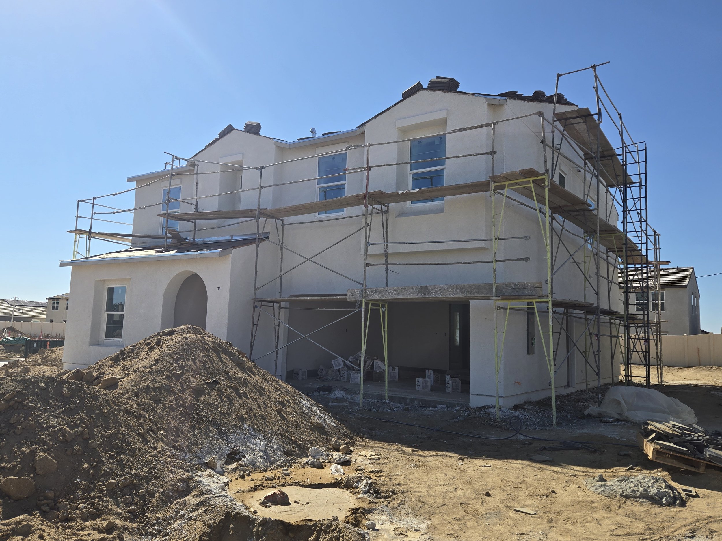 A large house under construction with scaffolding surrounding it, piles of dirt and construction debris in the foreground, and a clear blue sky.