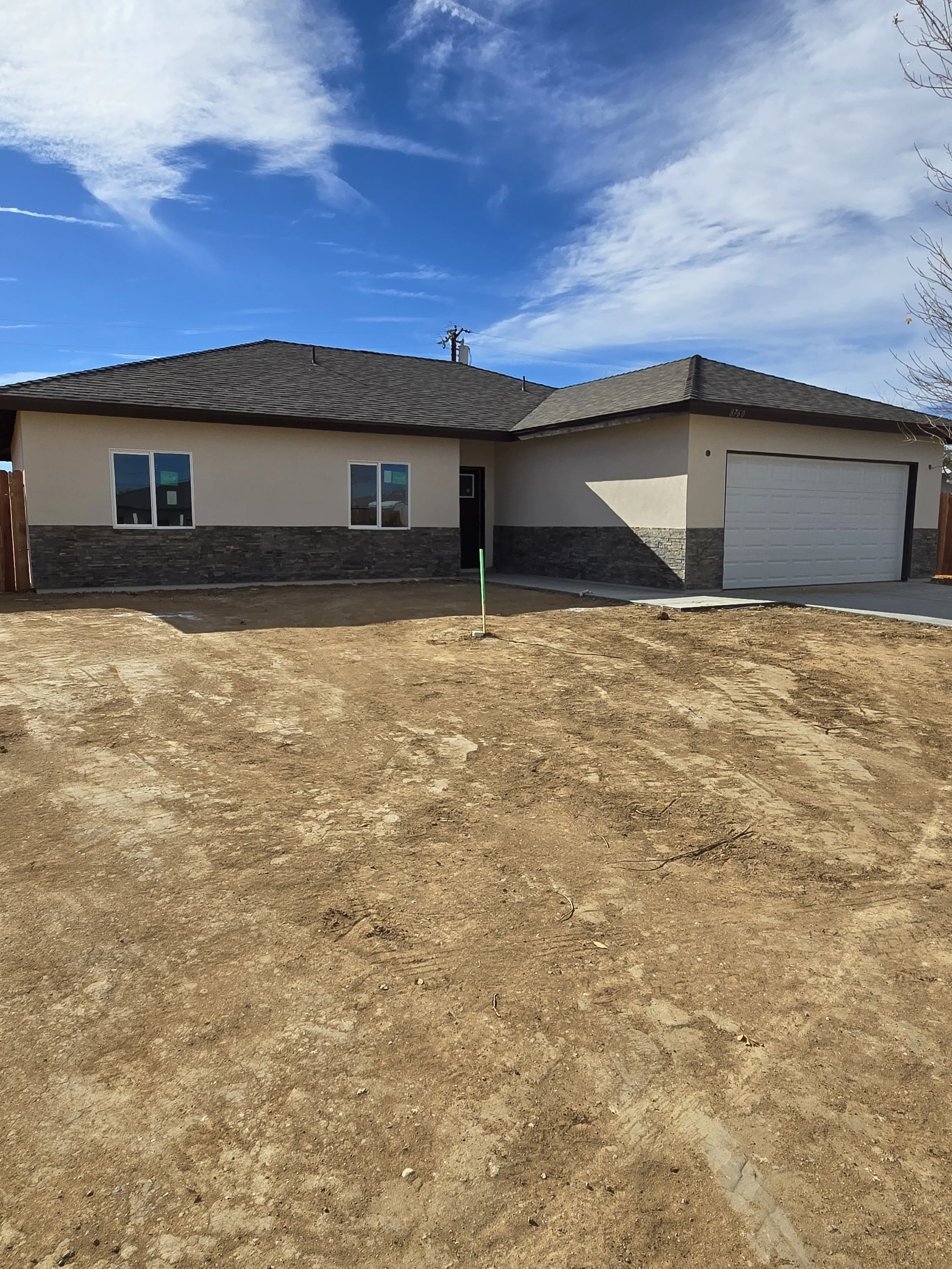 Newly constructed single-story house with gray and beige exterior, stone accents, two windows, a garage, and a dirt yard.
