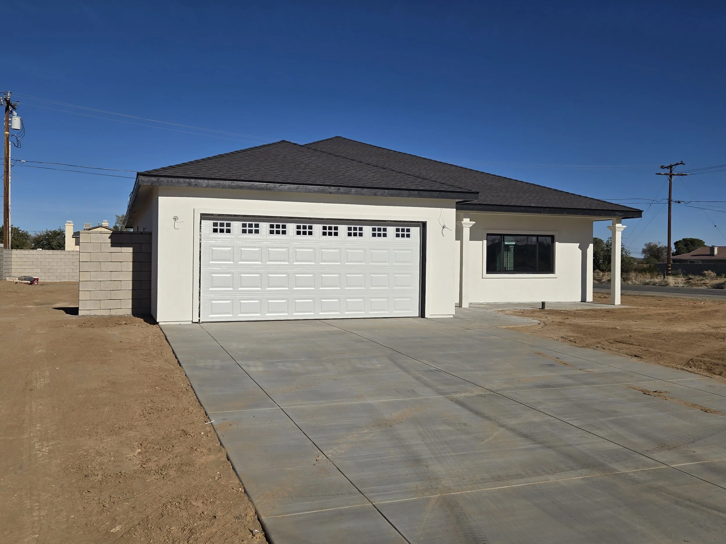 Newly constructed single-story house with a white exterior, gray shingle roof, large window, and attached garage with a white door. The house is situated in a developing area with a concrete driveway and bare dirt around the property.