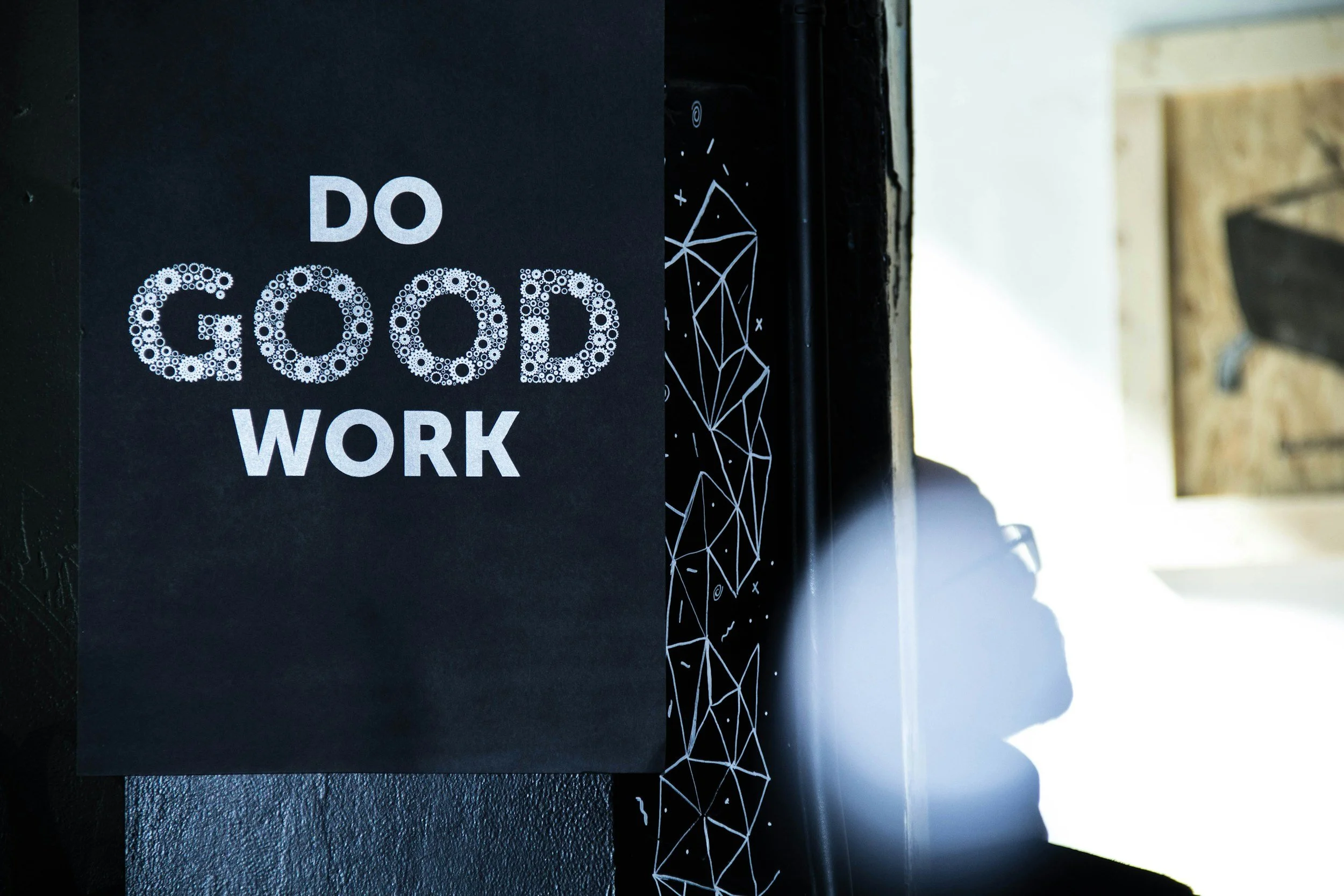 A black poster with white text that says 'Do Good Work,' with the word 'Good' made up of small white circles, attached to a black surface with a geometric white line pattern, light shines from the right side.