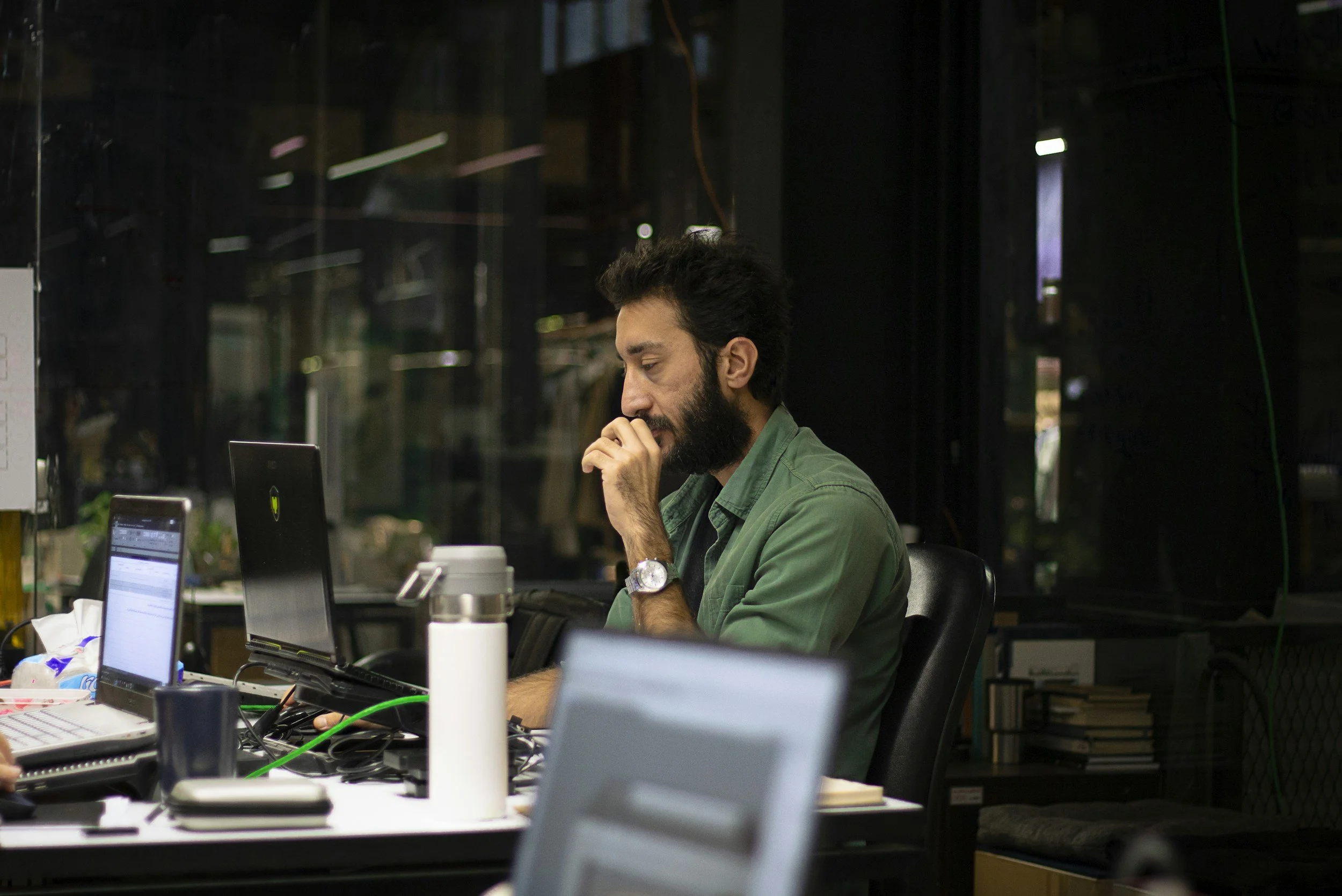 A man with dark hair and a beard, wearing a green shirt and a watch, sitting at a desk with multiple laptops and other items, looking focused with his hand near his mouth.