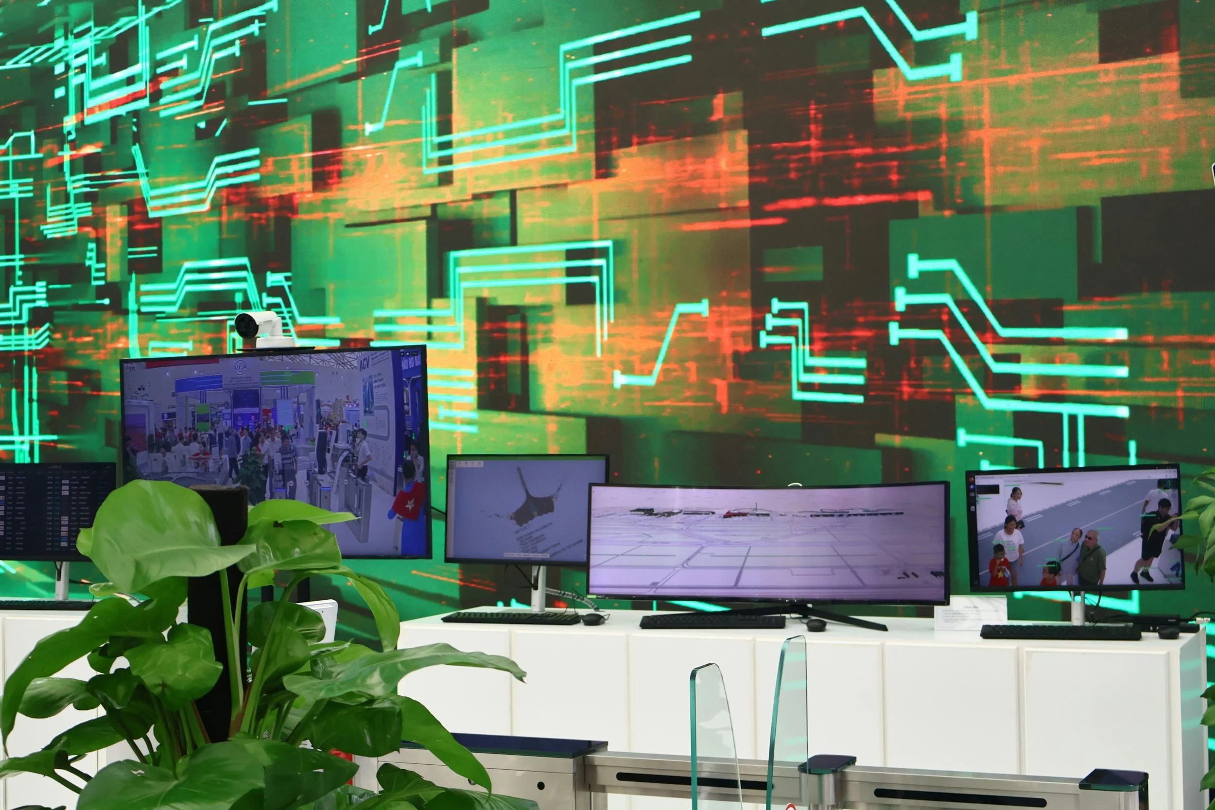 An indoor control room with multiple computer monitors displaying various scenes, including a crowded event, aircraft, and airport runway. A large digital screen with a colorful abstract circuit board pattern serves as the background, and there are green plants in the foreground.