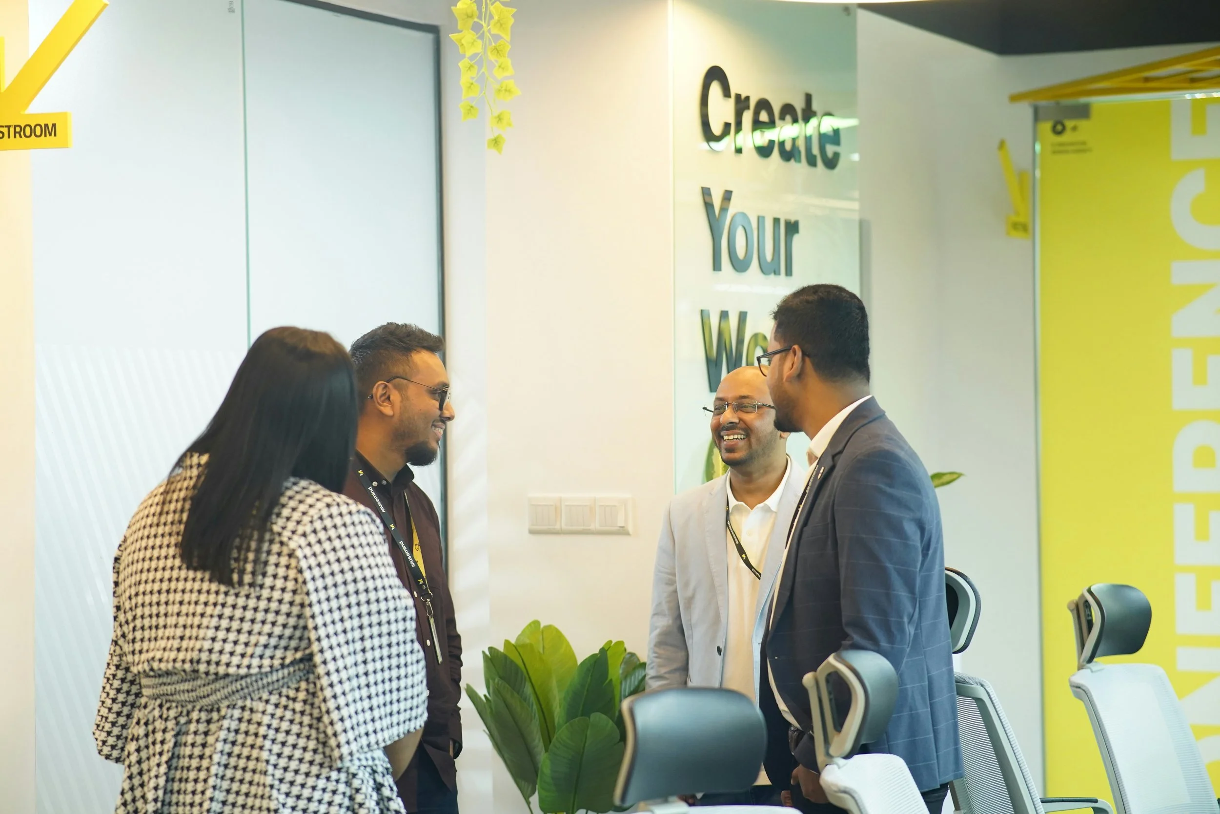 Four diverse professionals, two men and two women, are standing and talking in an office. They are smiling and appear to be engaged in a friendly conversation. The office has modern decor with yellow accents, plants, and a sign on the wall that says 