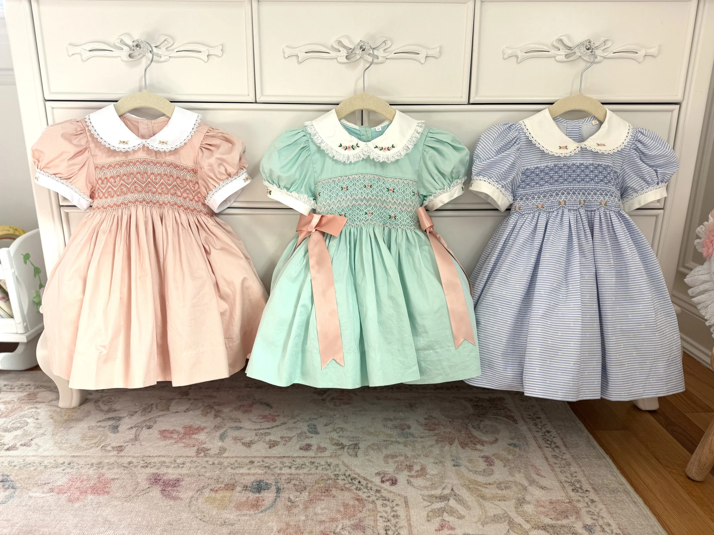 Three children's dresses in pastel colors hanging on a white cabinet.
