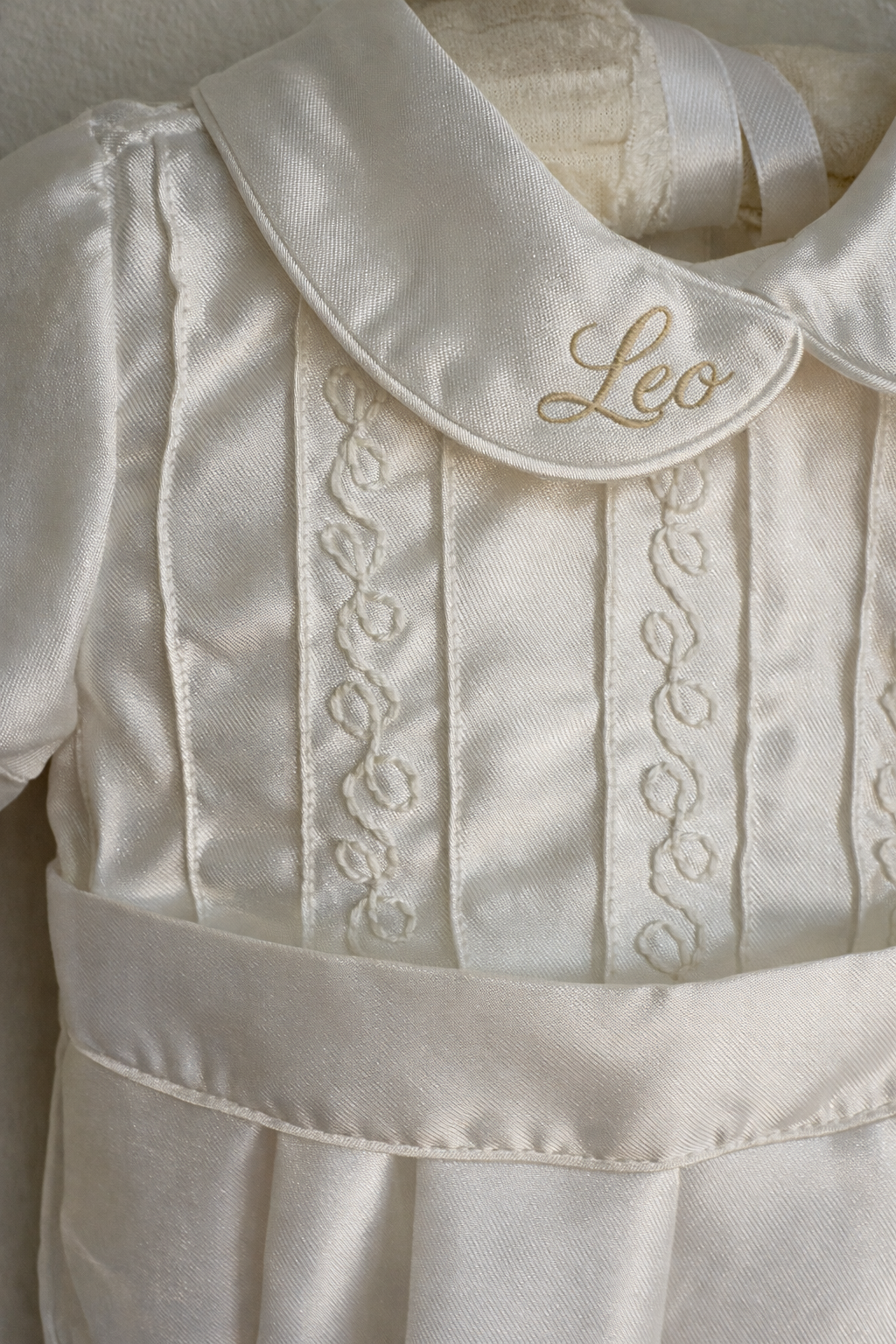 Close-up of a white satin christening gown with embroidered details and a collar embroidered with the name 'Leo' in gold.