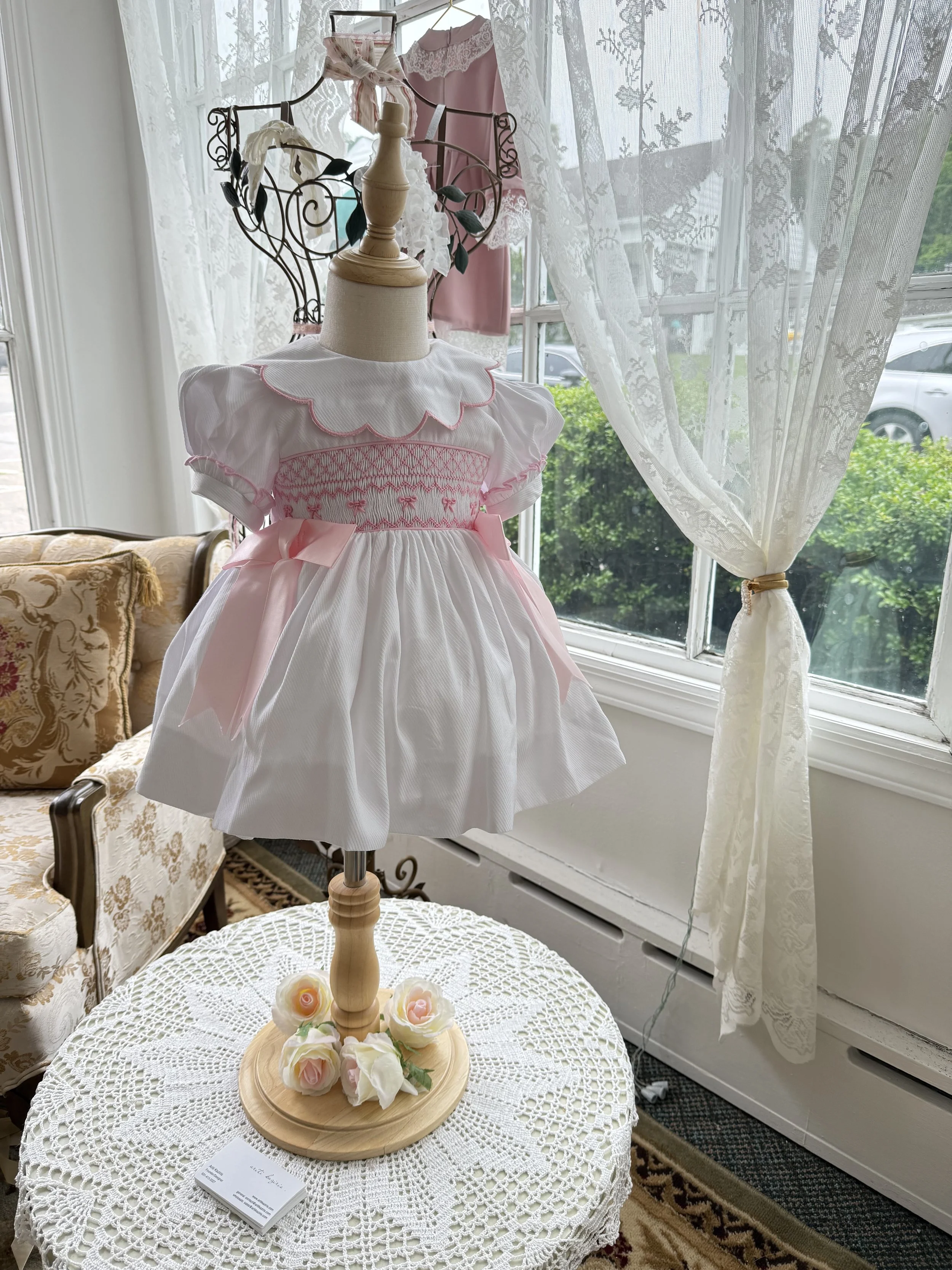 A white and pink baby dress displayed on a mannequin with a lace collar and pink bows, placed on a lace-covered table. In the background, there are lace curtains, an ornate vintage sofa, and a clothing rack with additional baby clothes.