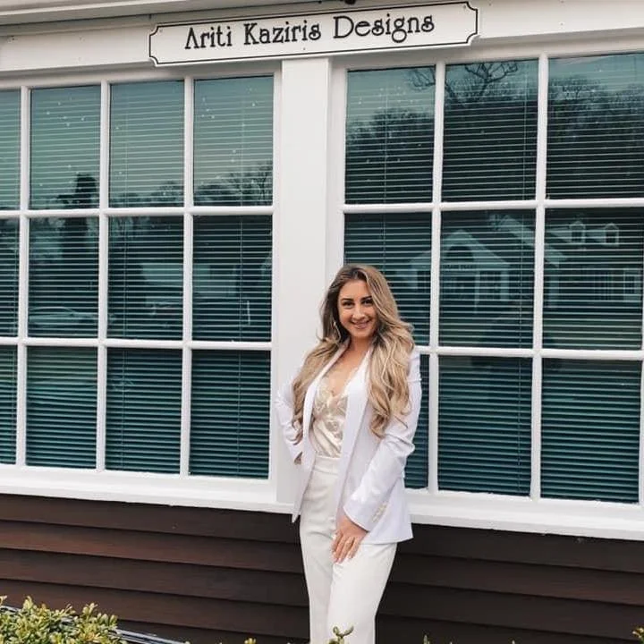 A woman with long blonde hair wearing a white suit standing outside a storefront with the sign 'Ariti Kaziris Designs' above the window.