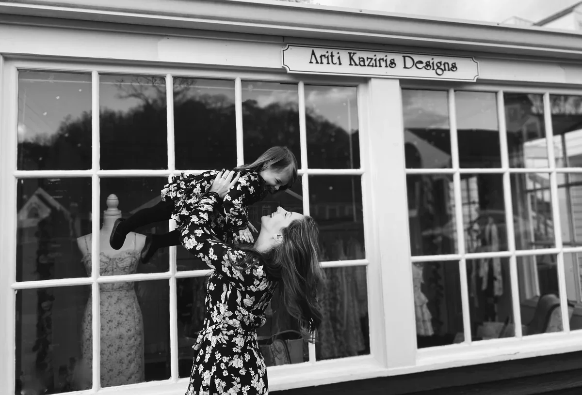 A woman and a young girl in floral dresses sharing a joyful moment outside a boutique store called 'Ariti Kaziris Designs'. The woman lifts the girl in the air, both smiling at each other.