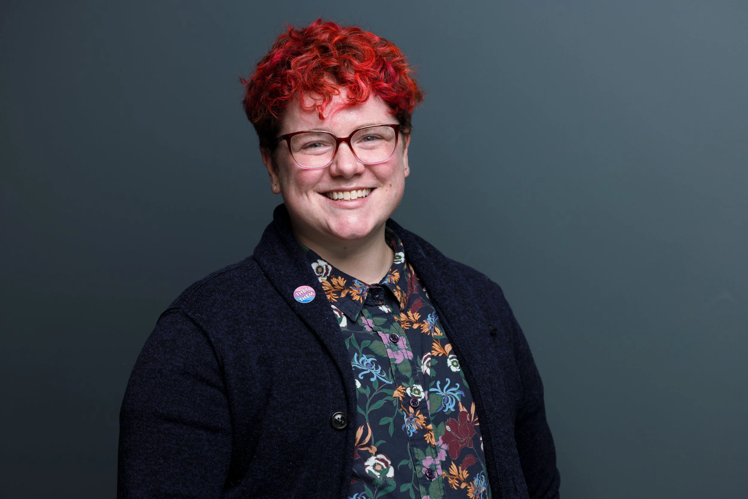 MM Petranoff, a person with dyed, bright red hair and classes. They wear a floral button-down and cardigan with a pin on their lapel that reads they/them.