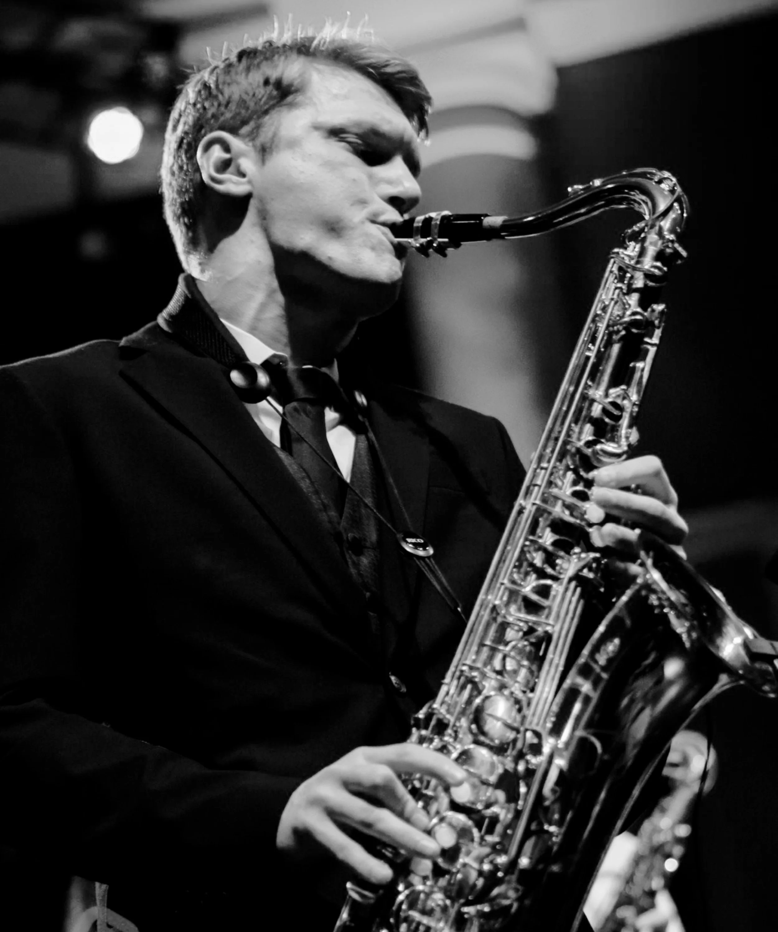 Black and white photo of a man playing a saxophone on stage, dressed in a suit with a bowtie.