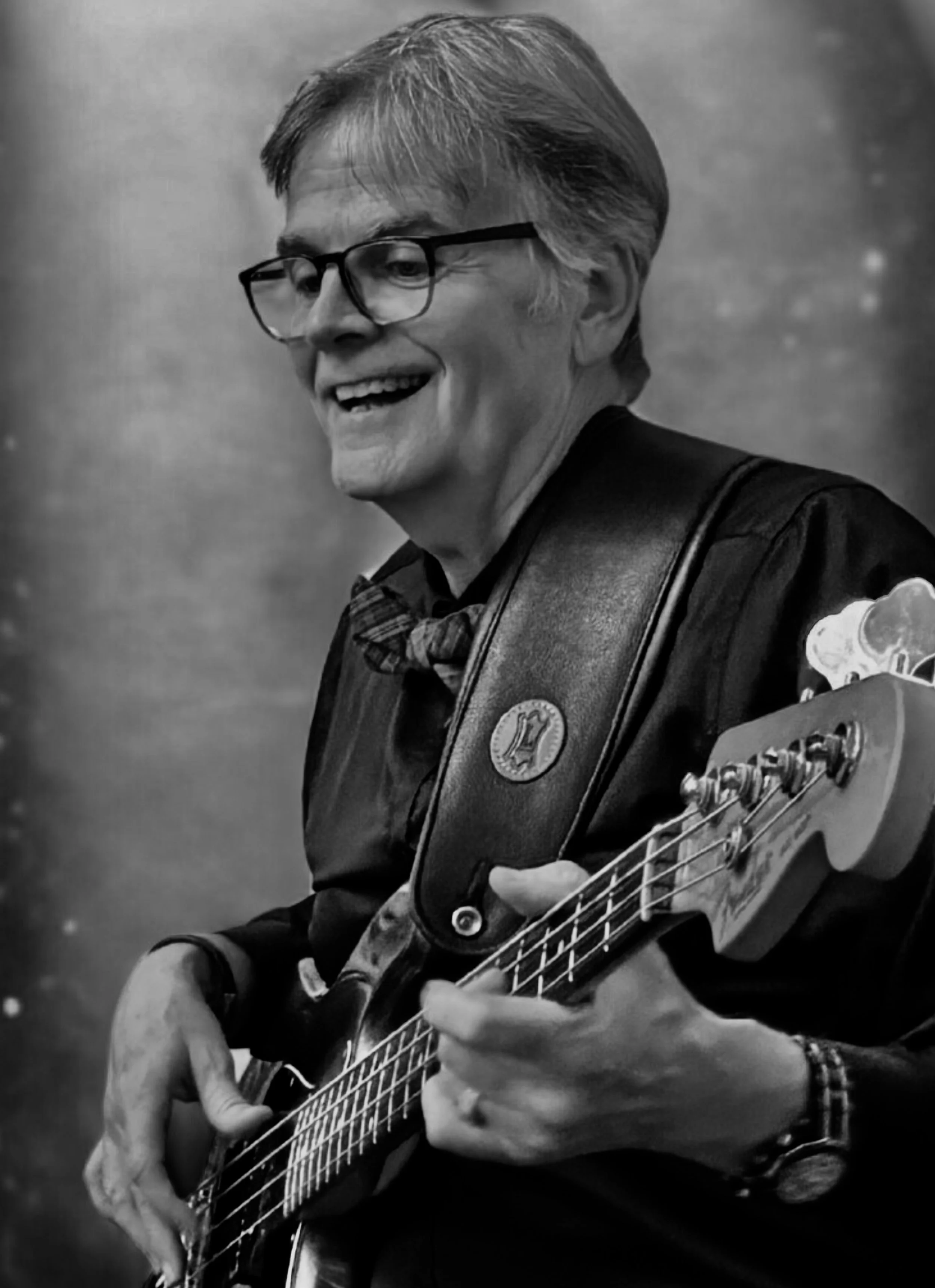 A black and white photo of a man with glasses smiling playing an electric bass guitar, wearing a leather jacket, a bowtie, and a wristwatch.
