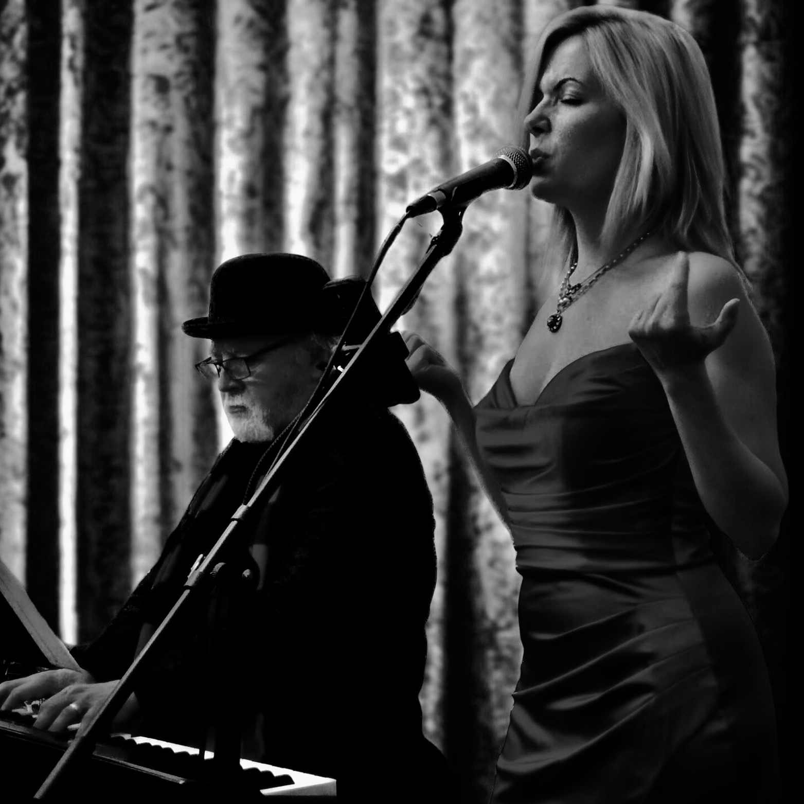 A woman singing or speaking into a microphone with a pianist playing in the background, set against a curtain backdrop, in a black and white photo.
