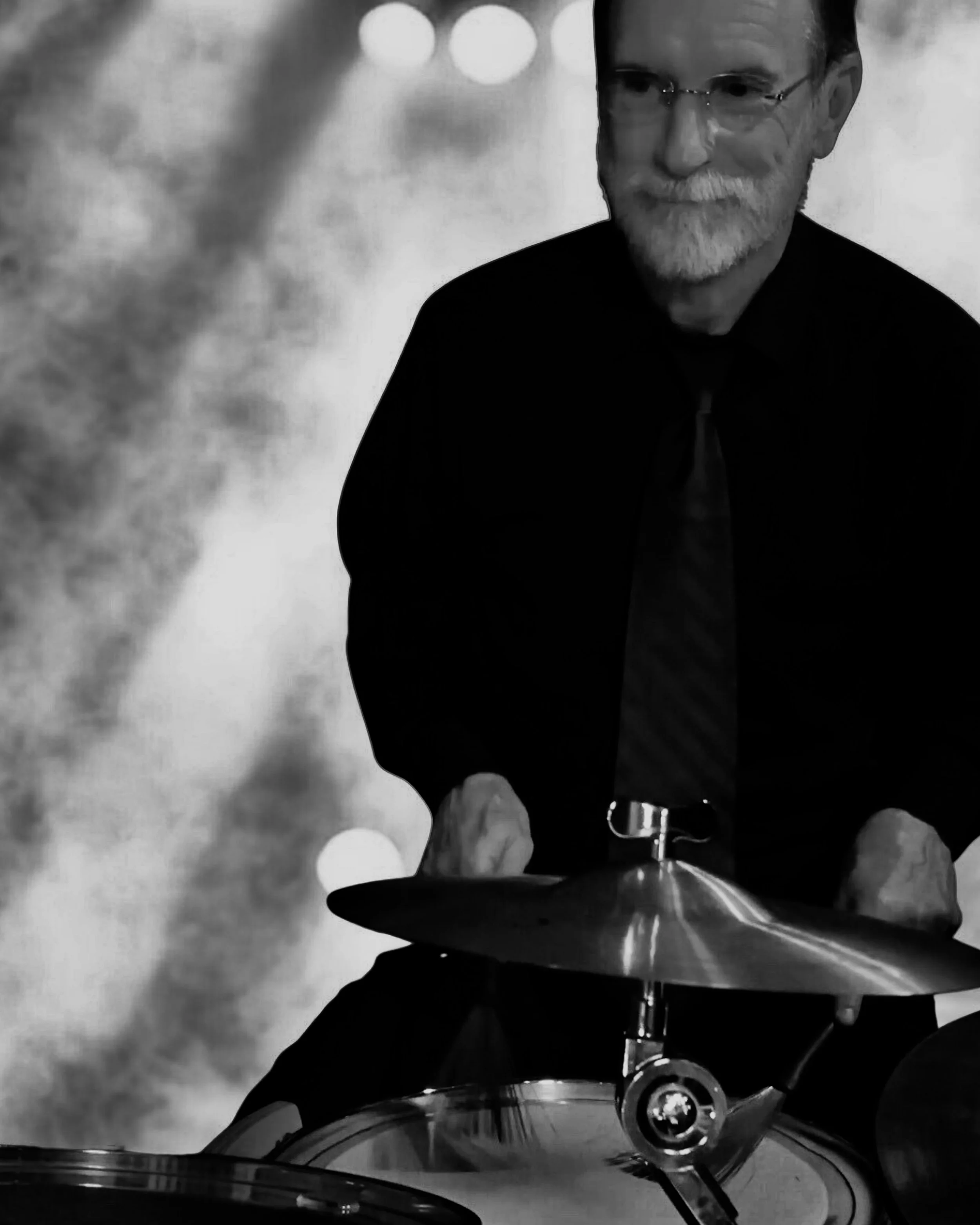 A man with glasses and a beard playing the drums, wearing a dark shirt and tie, in a performance setting with blurred lights in the background.
