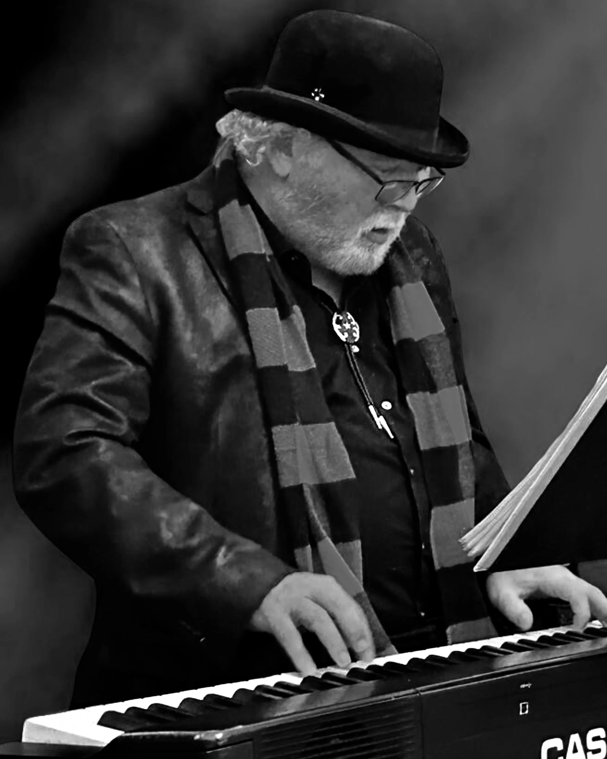 An elderly man with gray hair and beard playing a keyboard in black and white. He is wearing a black hat, glasses, a leather jacket, and a striped scarf, with sheet music in front of him.