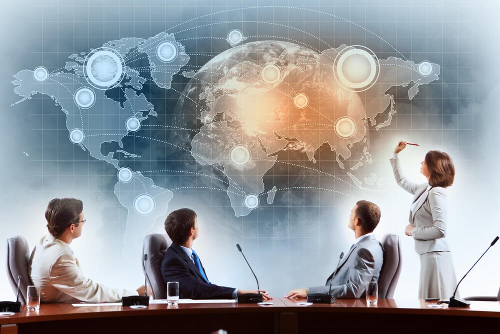 Business meeting with four people in a conference room, woman standing and presenting a digital world map with interconnected points projected on the wall.