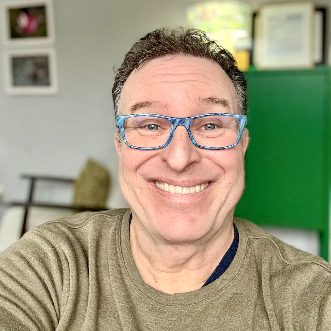 A smiling man wearing blue glasses and a brown shirt, taking a selfie indoors with framed photos and a green background in the room.