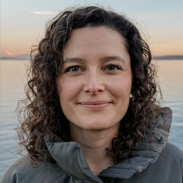 Close-up of a woman with curly brown hair, smiling outdoors near a body of water during sunset, wearing a gray jacket.