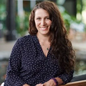 A woman with long, curly dark hair smiling, wearing a dark polka dot shirt, outdoors with greenery in the background.