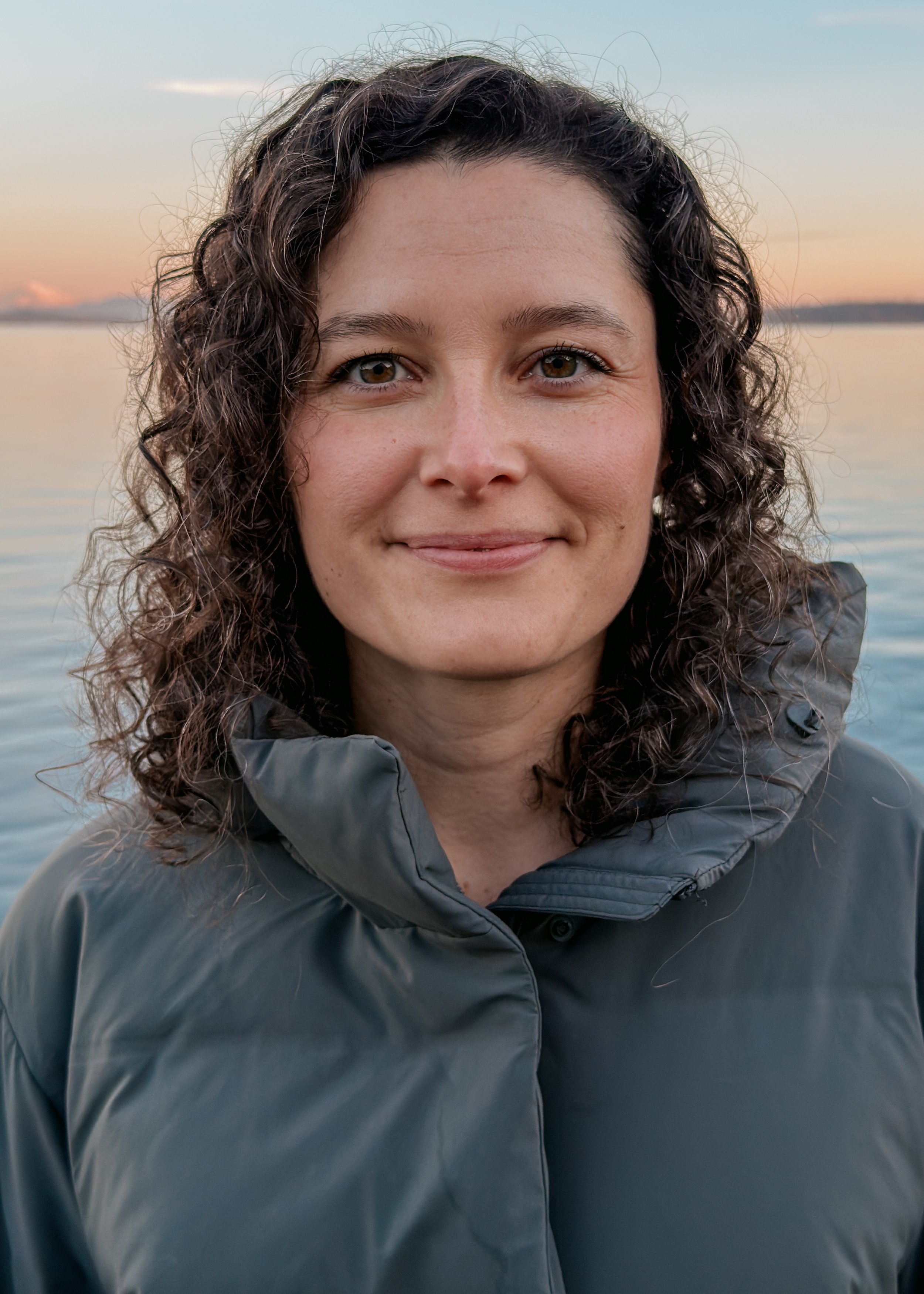 A woman with curly brown hair smiling outdoors near a body of water during sunset, wearing a gray jacket.