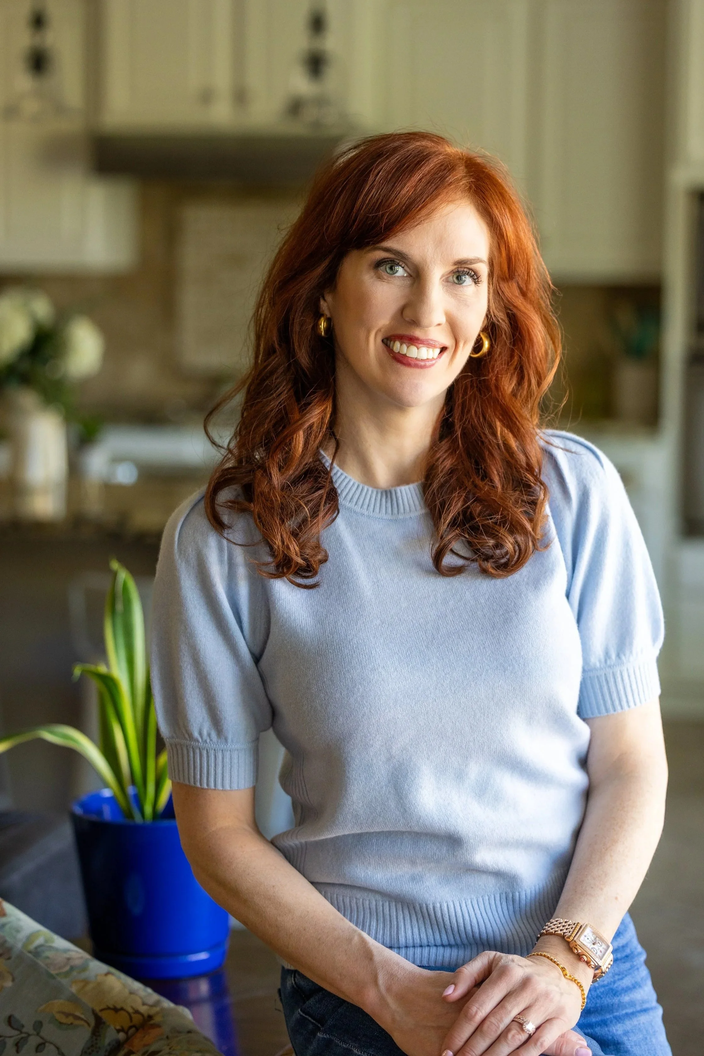 A woman with red hair smiling, wearing a light blue sweater and jewelry, sitting indoors with a background of a kitchen and a potted plant.