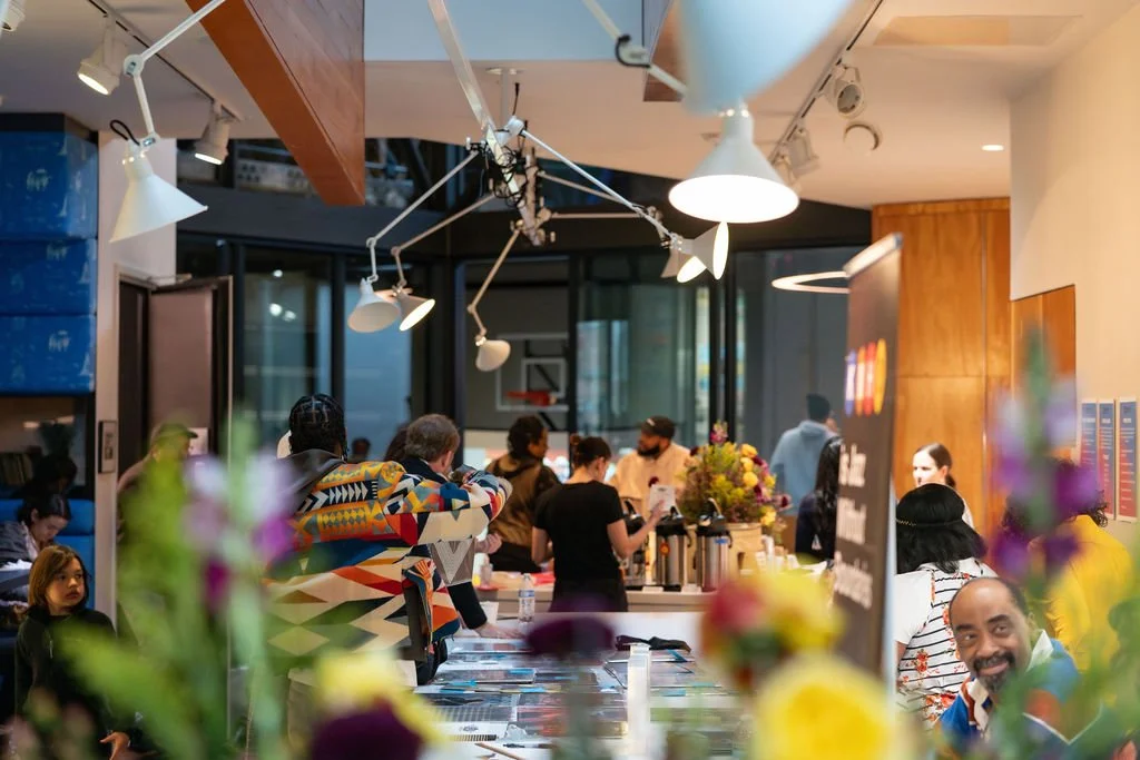 People waiting in line at a modern cafe or restaurant with floral arrangements and large windows.