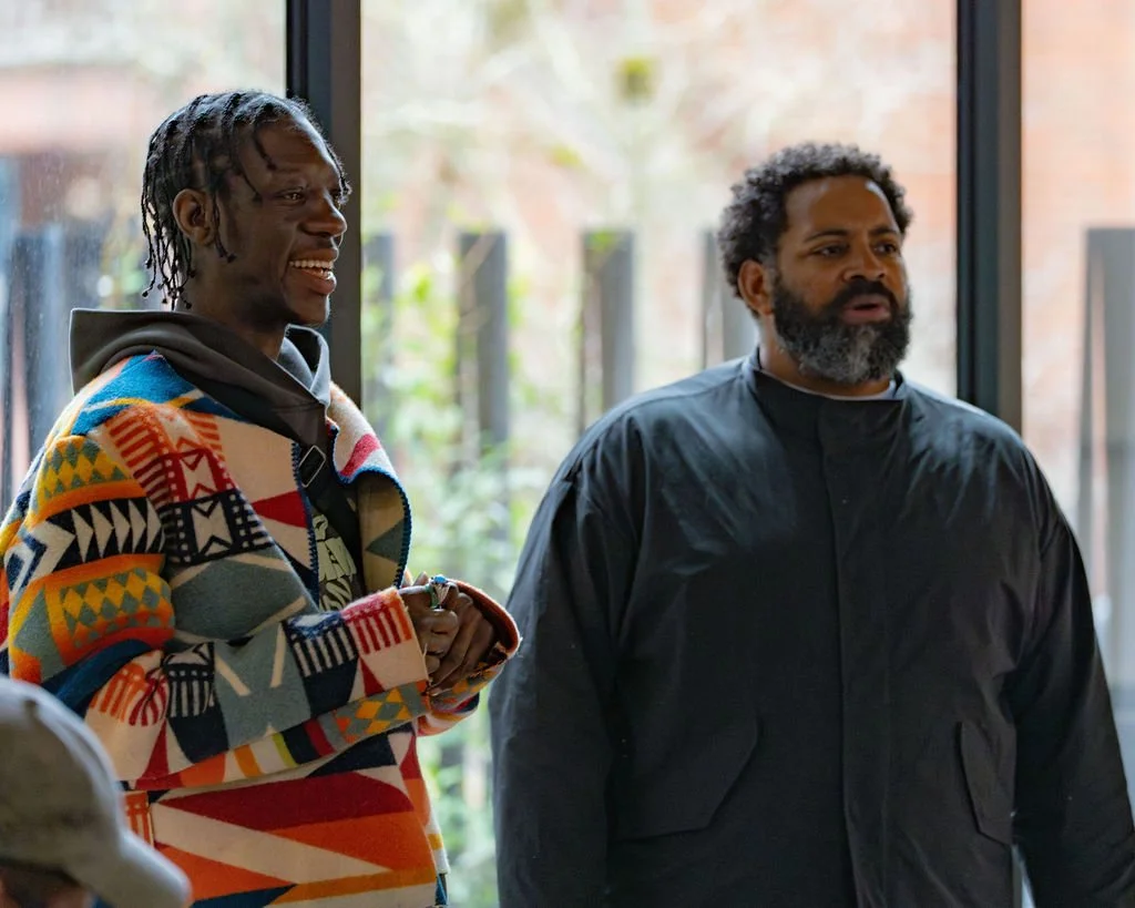 Two men standing indoors near large windows, having a conversation with natural light coming through. One man wears a colorful patterned jacket and the other wears a black jacket with a beard and curly hair.