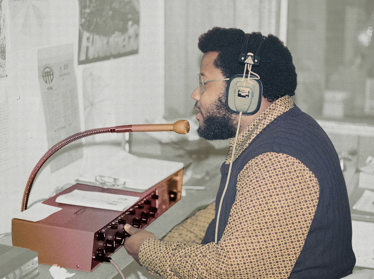 A man with curly hair and glasses wearing headphones, singing into a microphone and operating music equipment.