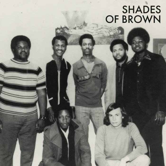 Black and white photo of five men standing and one woman sitting in front of them. The men are wearing casual shirts and jackets, and the woman has shoulder-length hair and is wearing a light-colored sweater. The background features a wall with an image of a donkey and the text "SHADES OF BROWN."
