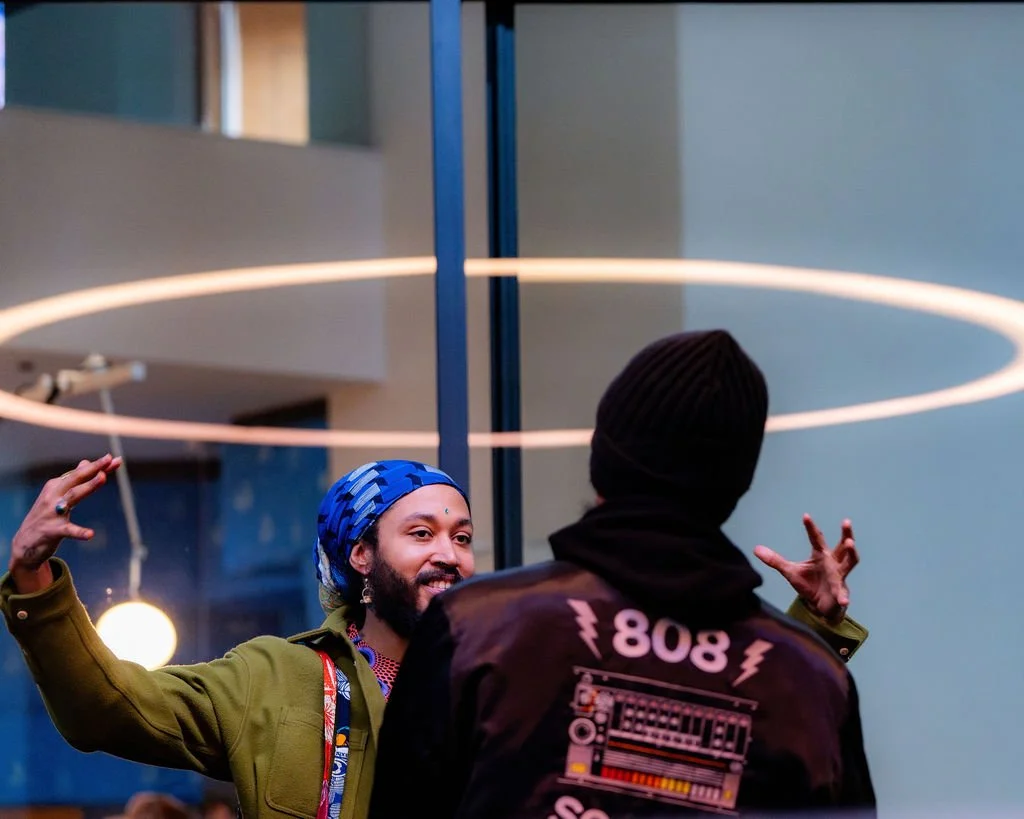 Two men are engaged in a conversation inside a modern building. One man, with a beard and colorful headscarf, is smiling and gesturing with his hands. The other man, wearing a black beanie and jacket with electronic music graphics, has his back to the camera. A circular light fixture is visible above them.