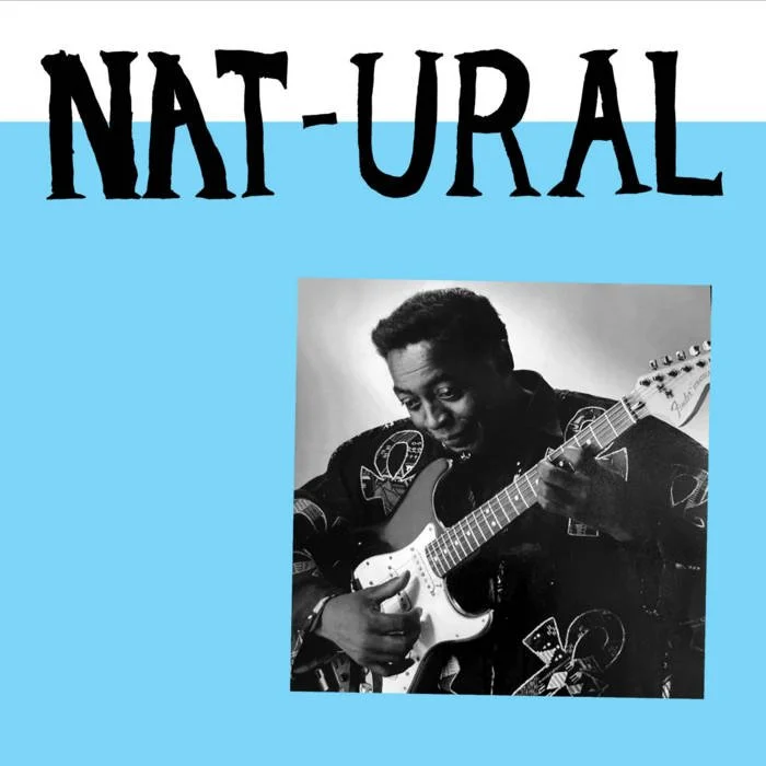 A black-and-white photo of a man playing an electric guitar. The background of the image is part of a cover with the text 'NAT-URAL' in large black letters on a blue background.