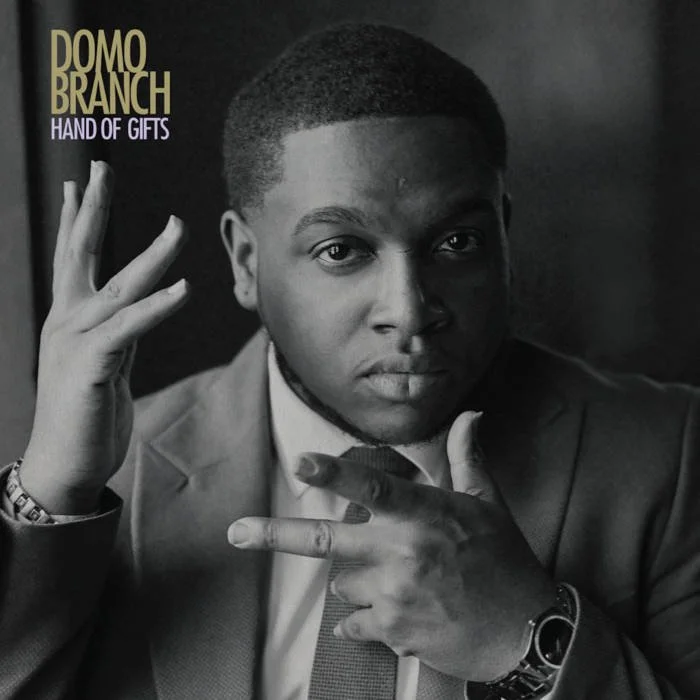 Black and white album cover featuring a man in a suit with a serious expression, holding his hands up near his face. Text in the upper left corner reads 'Domo Branch Hand of Gifts.'