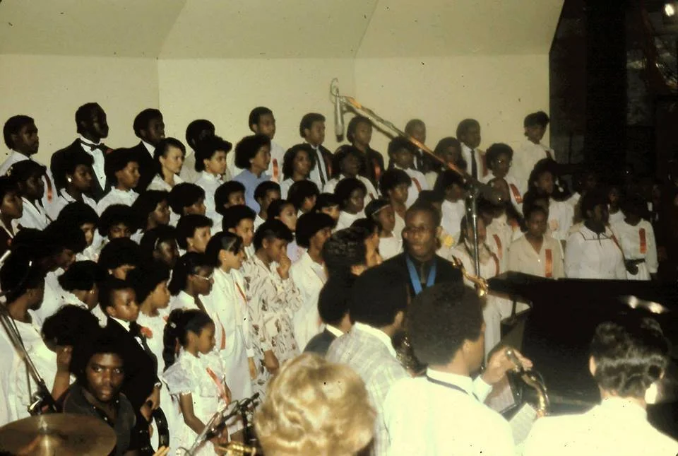 A large group of children singing or performing on stage, with some musicians playing instruments in front of them, in an indoor setting.