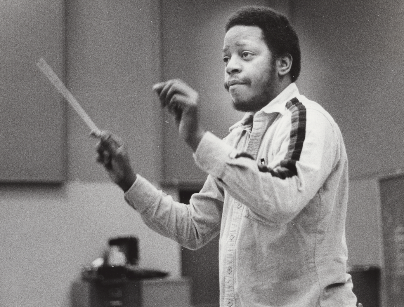 A black and white photo of a man with an afro hairstyle, wearing a light-colored jacket with dark stripes on the sleeves, gesturing with his hands while speaking or conducting in a room.