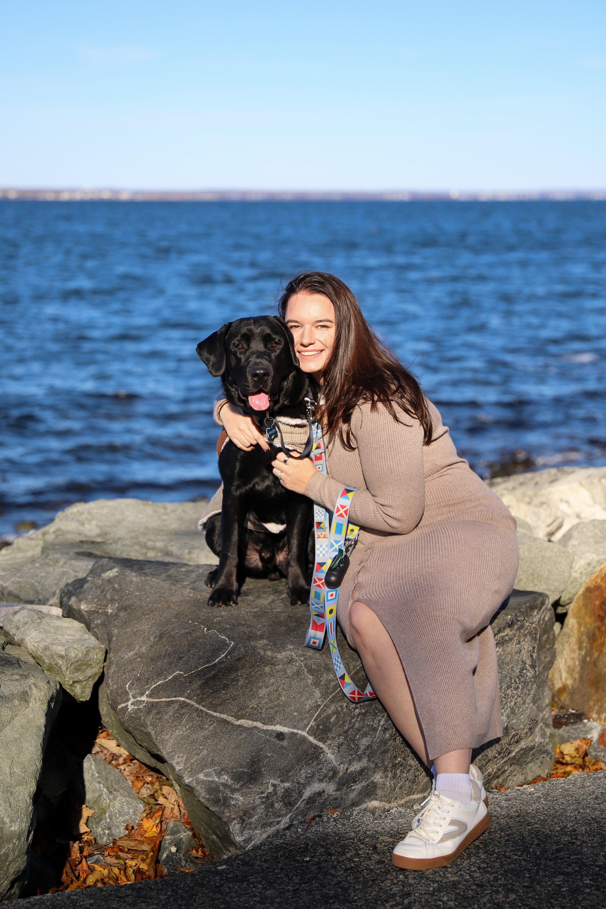 A woman with long brown hair wearing a beige dress and white sneakers, sitting on rocks by the water, hugging a black Labrador Retriever dog.