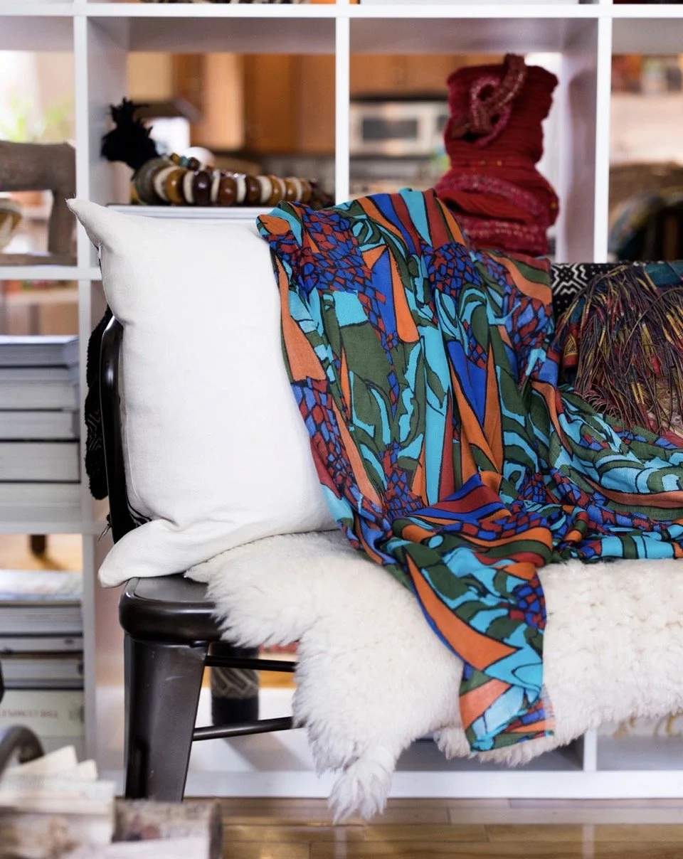 A cozy living room scene with a black chair covered with a fluffy white throw blanket, a white pillow, colorful patterned scarves and blankets draped over the chair, a bookshelf with a bead necklace and a red woven object on top, and a bookshelf in the background.