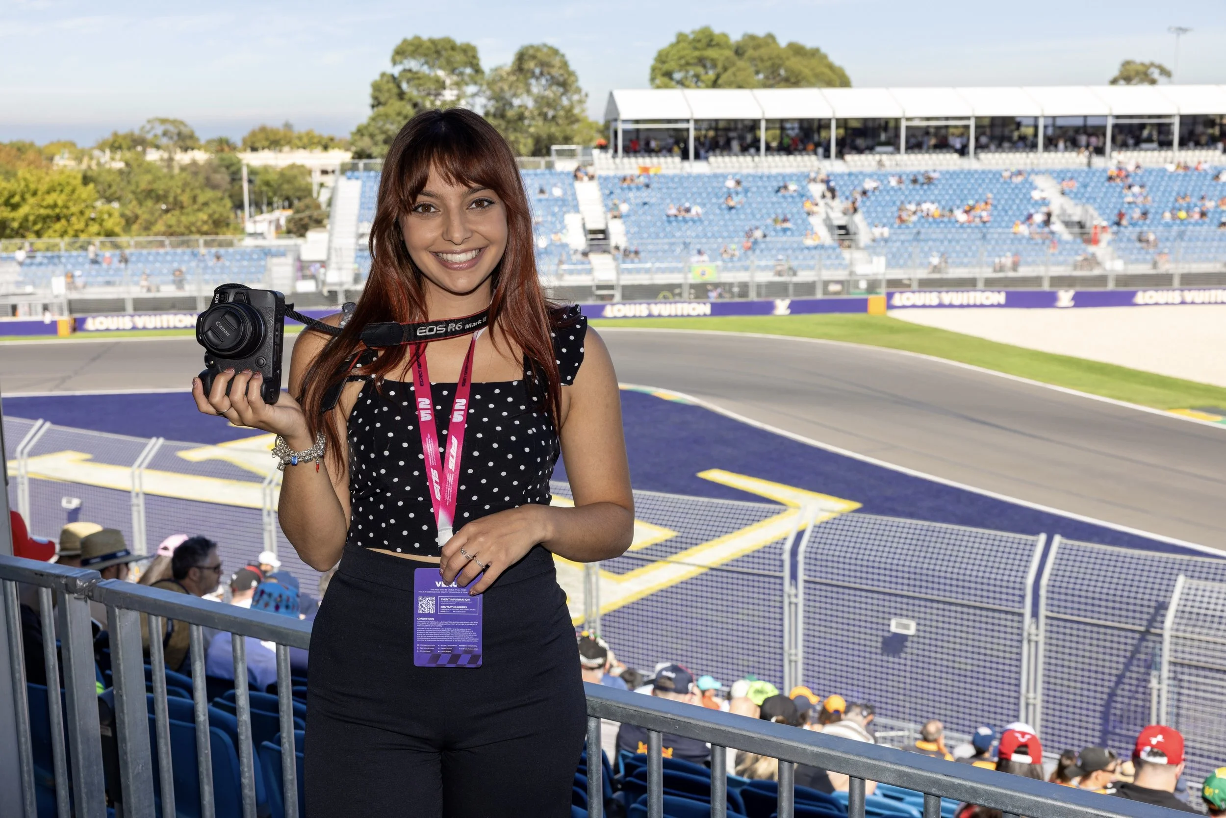 A woman smiling at a race track, holding a camera with a strap around her neck. She is wearing a polka dot top with black pants, and an event badge around her neck. The background shows a race track with spectators in the stands.
