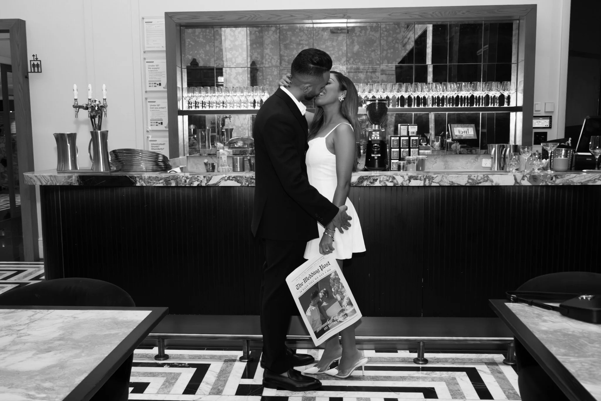 A couple, dressed elegantly, sharing an intimate moment behind a bar counter in a stylish restaurant, with the man holding a newspaper.