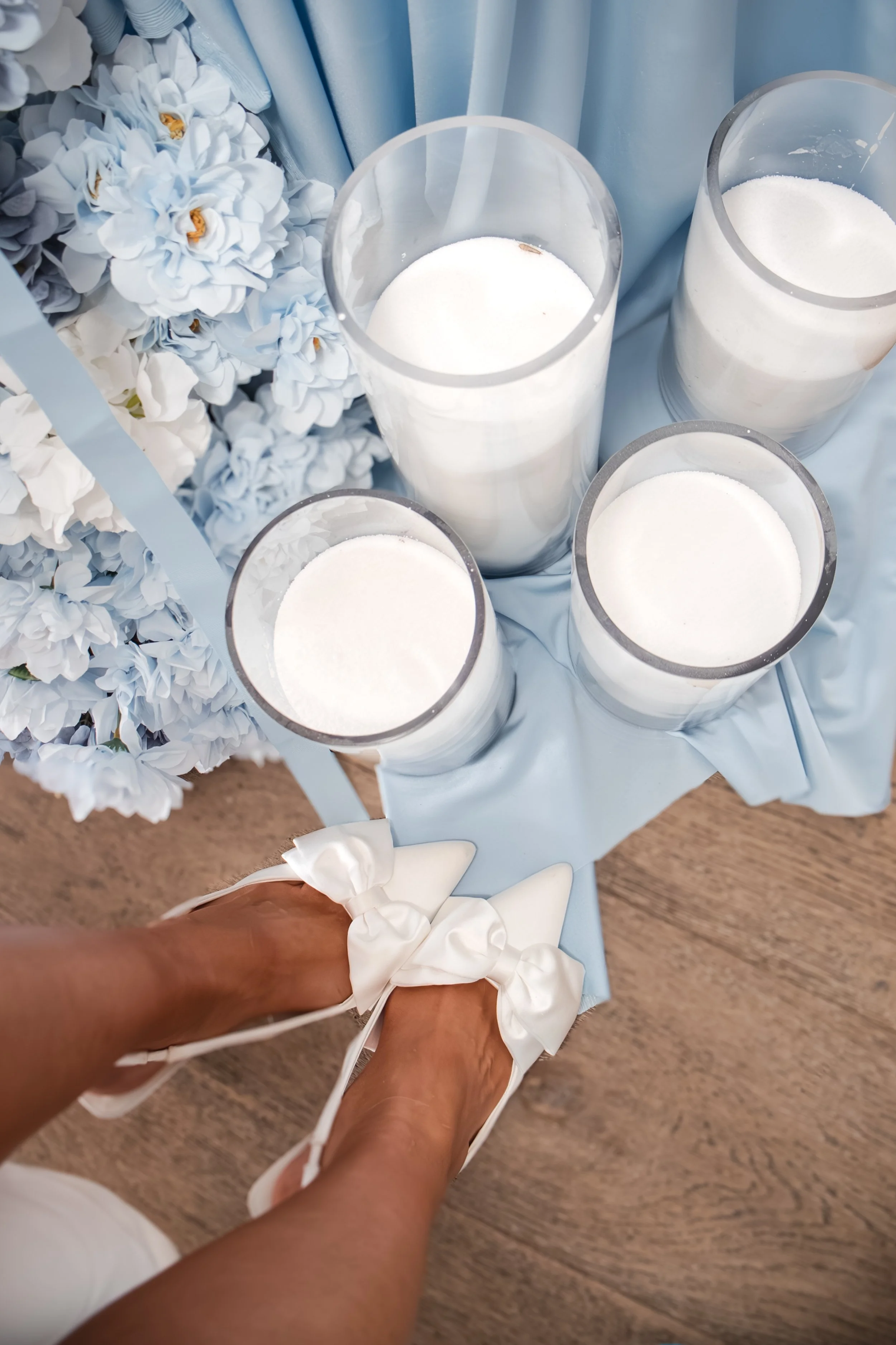 Four white candles in glass holders placed on a light blue cloth on a wooden surface, with blue and white flowers nearby.