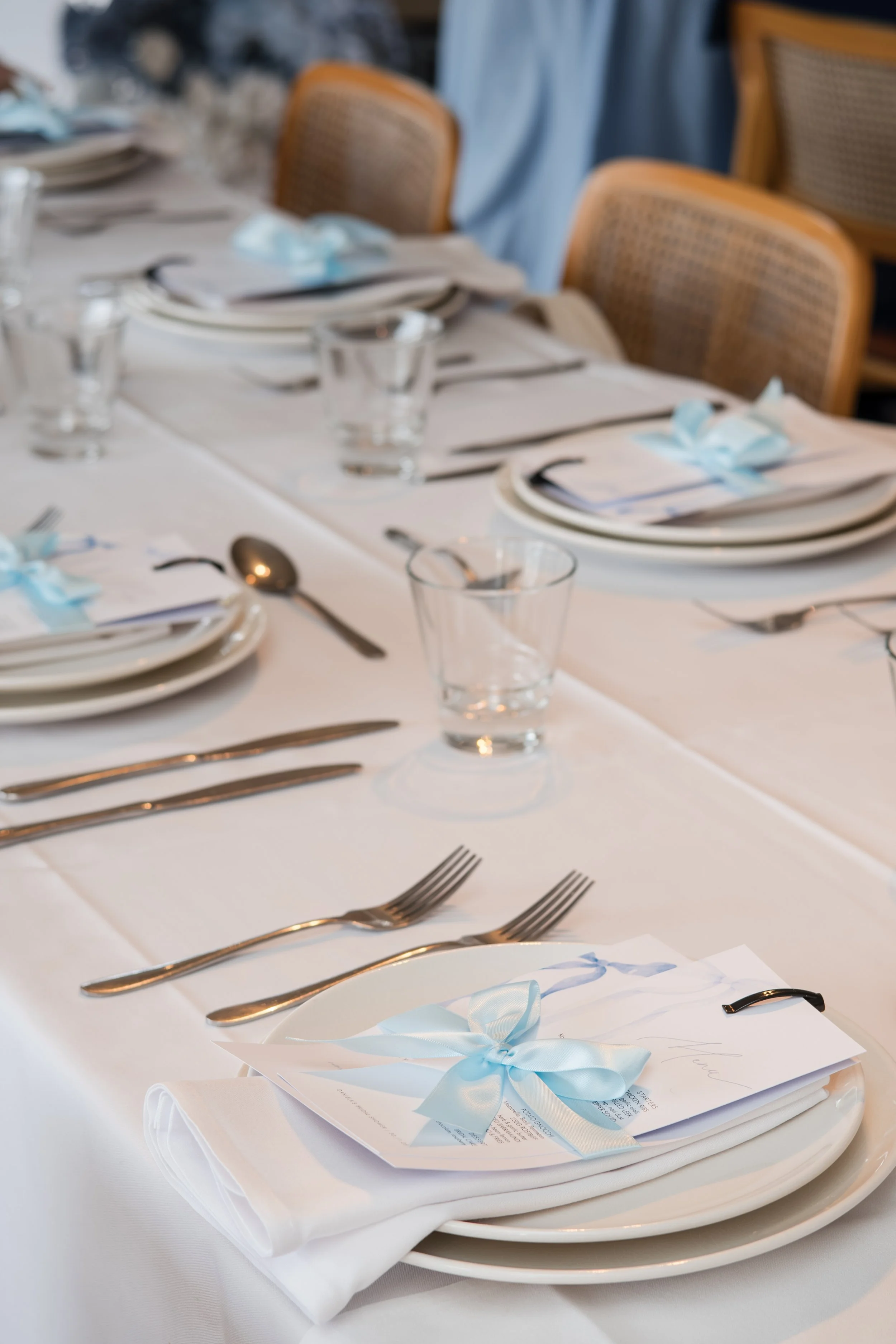 A formal dining table set with white tablecloth, plates, silverware, glasses, and napkins with blue ribbons and cards for a special occasion.