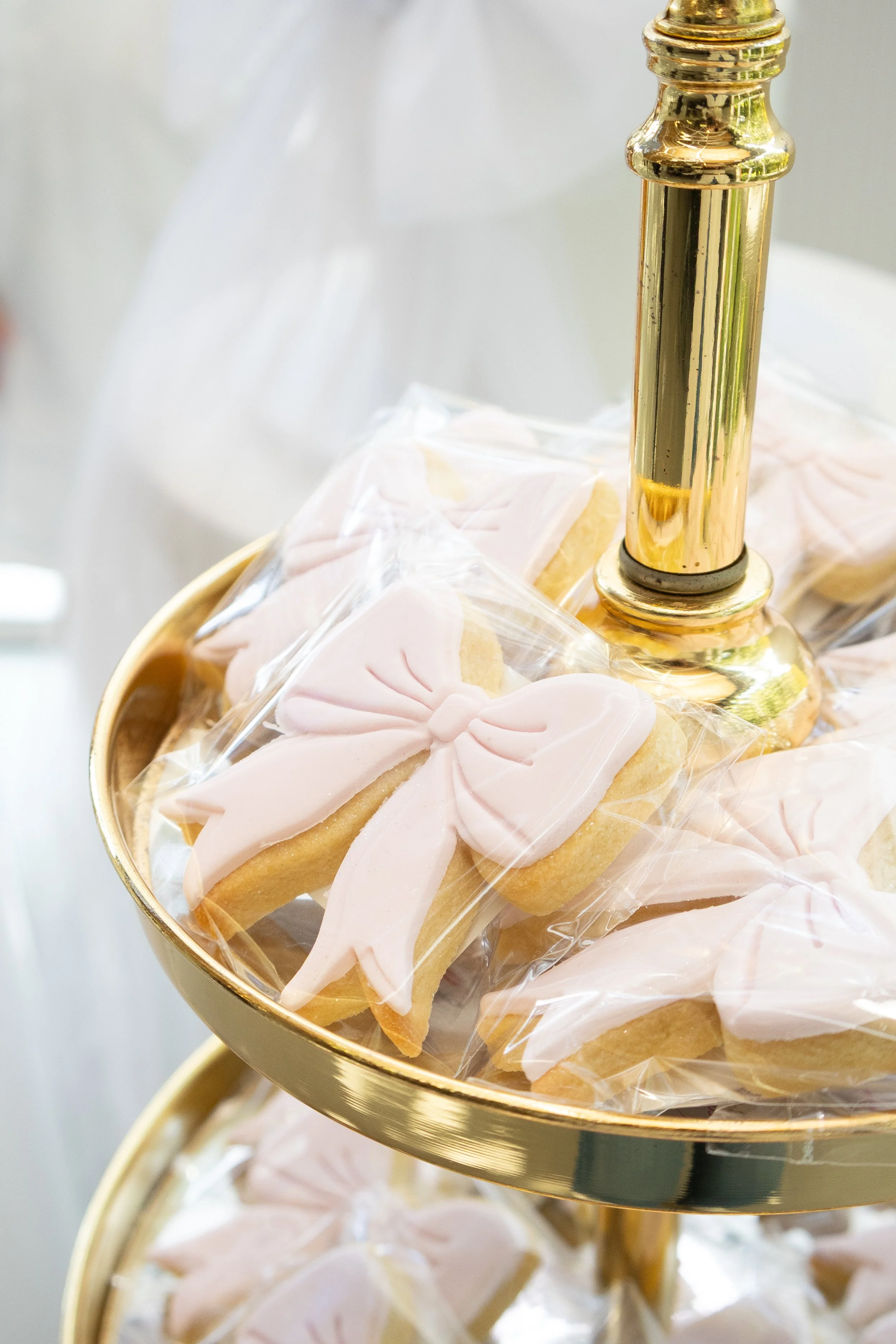 Decorative cookies with pink icing in the shape of bows, displayed on a gold and white tiered stand.